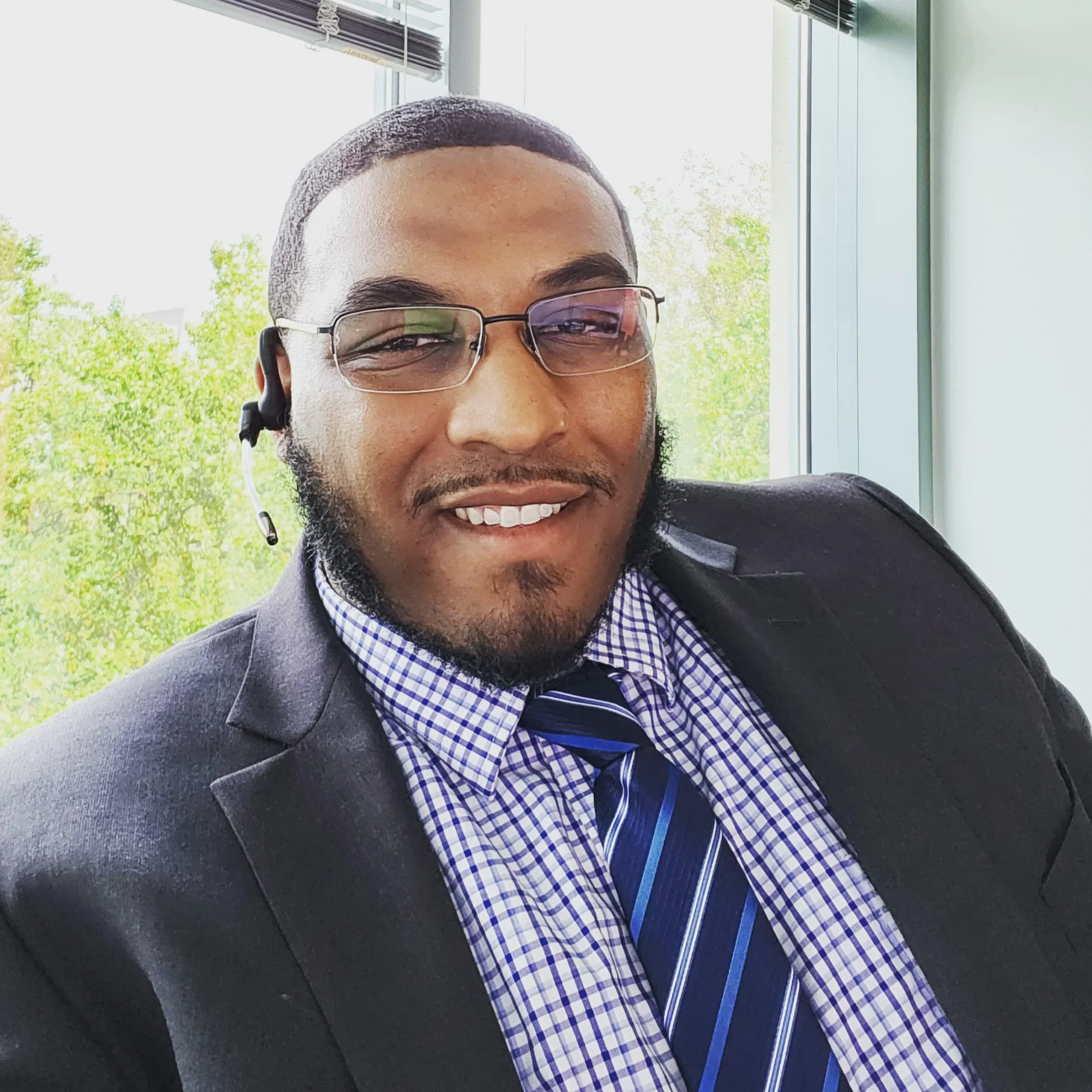 A smiling man in formal attire, wearing a suit, dress shirt, and striped tie, sitting near a window with a suburban or park view of green trees, and wearing glasses and an earpiece.