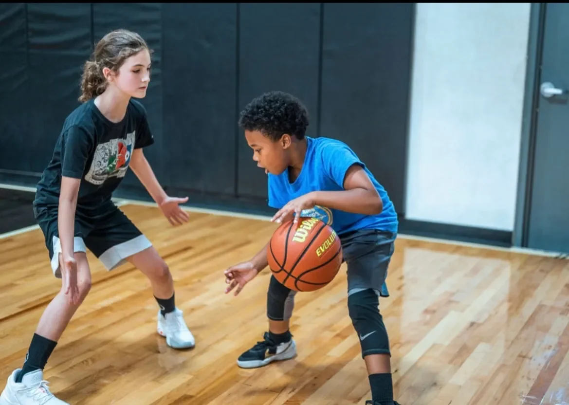 Two children playing basketball indoors on a wooden court, with one girl defending and one boy dribbling a basketball.