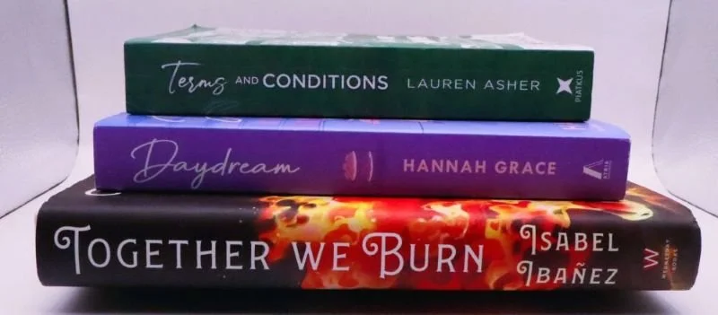 Stack of romance and fantasy books by Lauren Asher, Hannah Grace, and Isabel Ibañez with floral reader bookmark