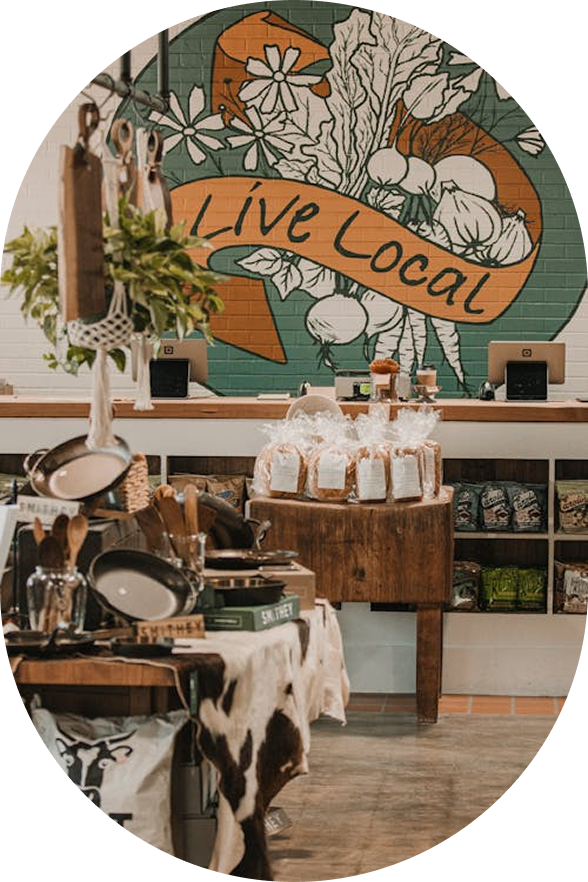 Interior of a store with a colorful mural on the wall that reads 'Live Local' and features vegetables and flowers. The store has various kitchen utensils, small items, and packaged goods displayed on tables and shelves.