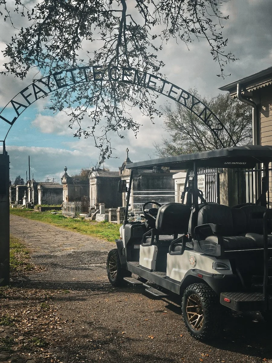 A golf cart parked near a cemetery entrance during the daytime, with cranes and tombstones visible in the background, and a sign reading 'Layfayette Cemetery No. 1' arching over the scene.