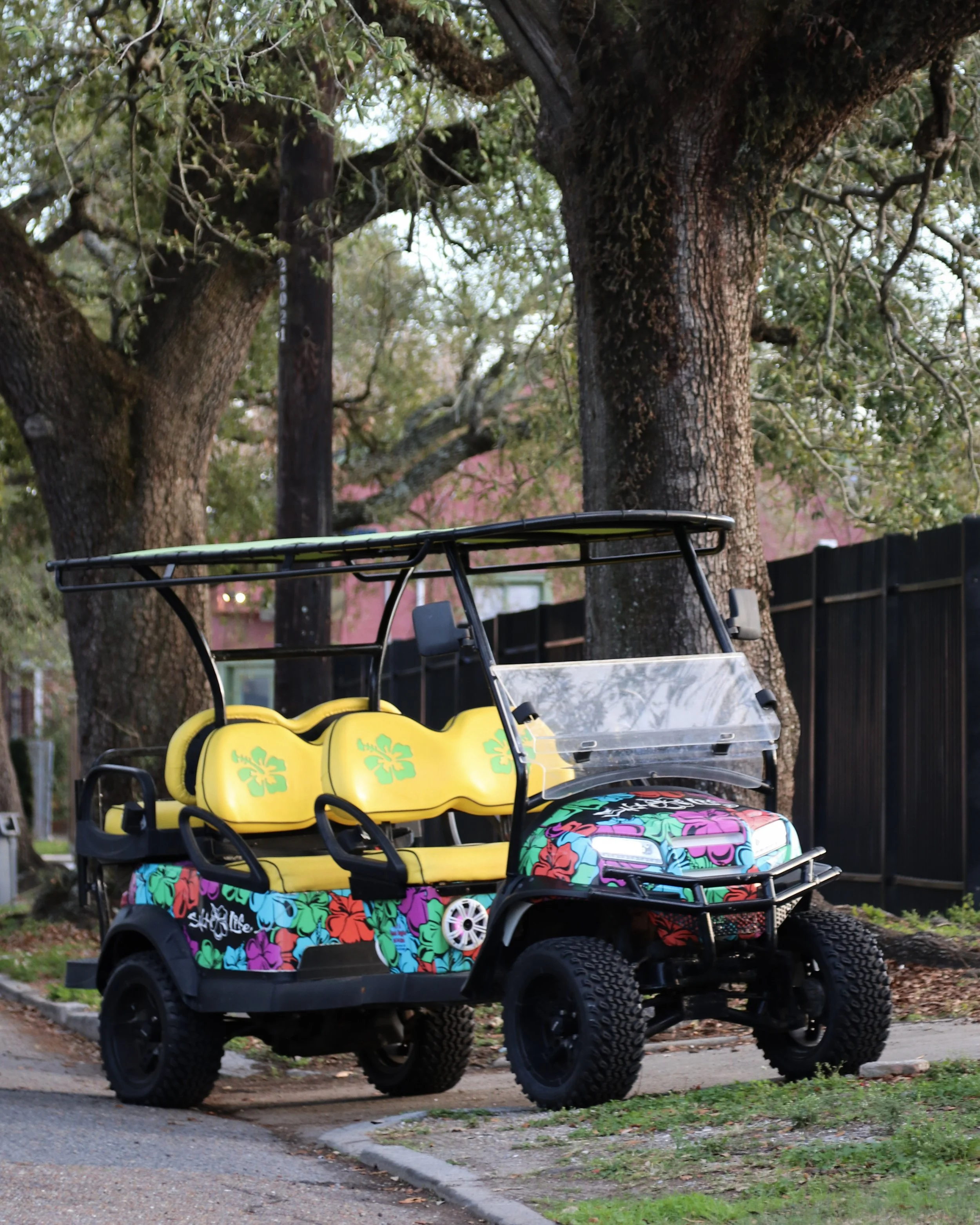 Colorful golf cart with yellow and floral-patterned seats parked on a sidewalk near trees and a black fence.