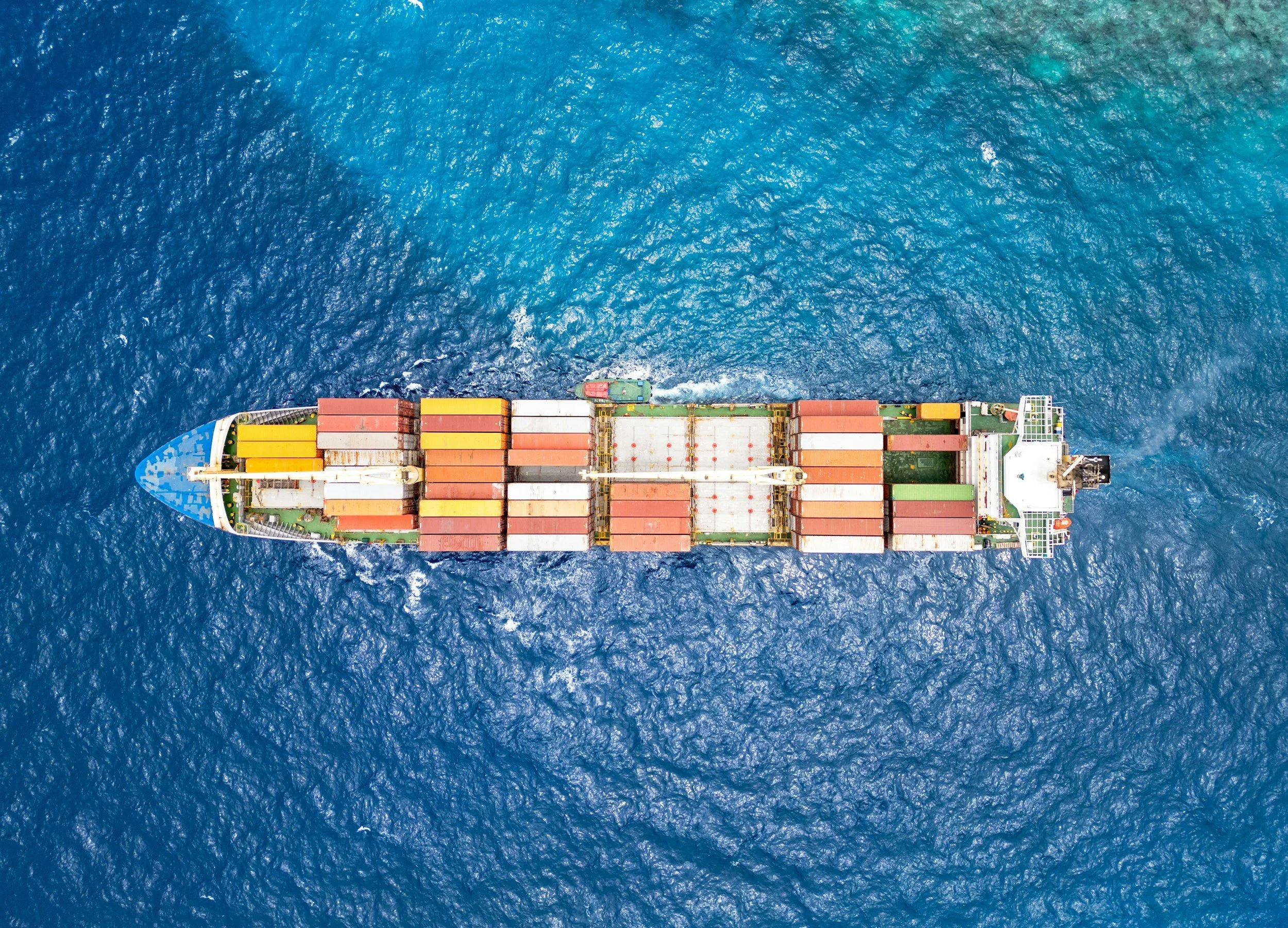 An aerial view of a cargo ship sailing on the ocean, carrying numerous colorful containers on deck.