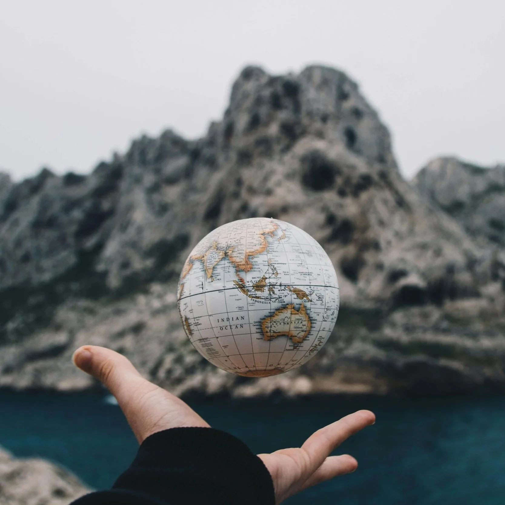 Person holding a mini globe with Asia and Australia visible, in front of rocky cliffs and ocean.