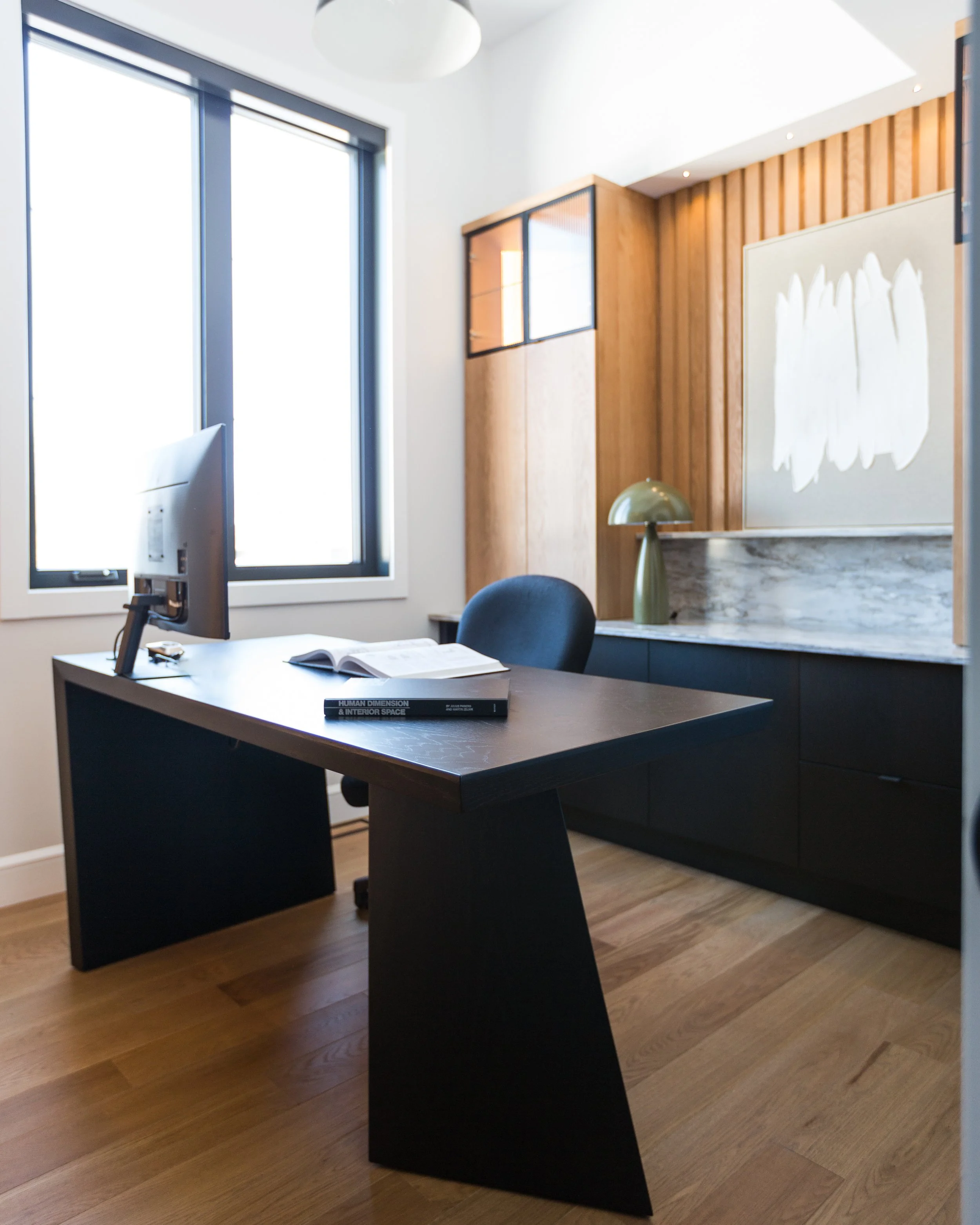 Modern office with a wooden accent wall, black desk, chair, and a computer, next to a large window with white blinds and a decorative lamp on the cabinet.