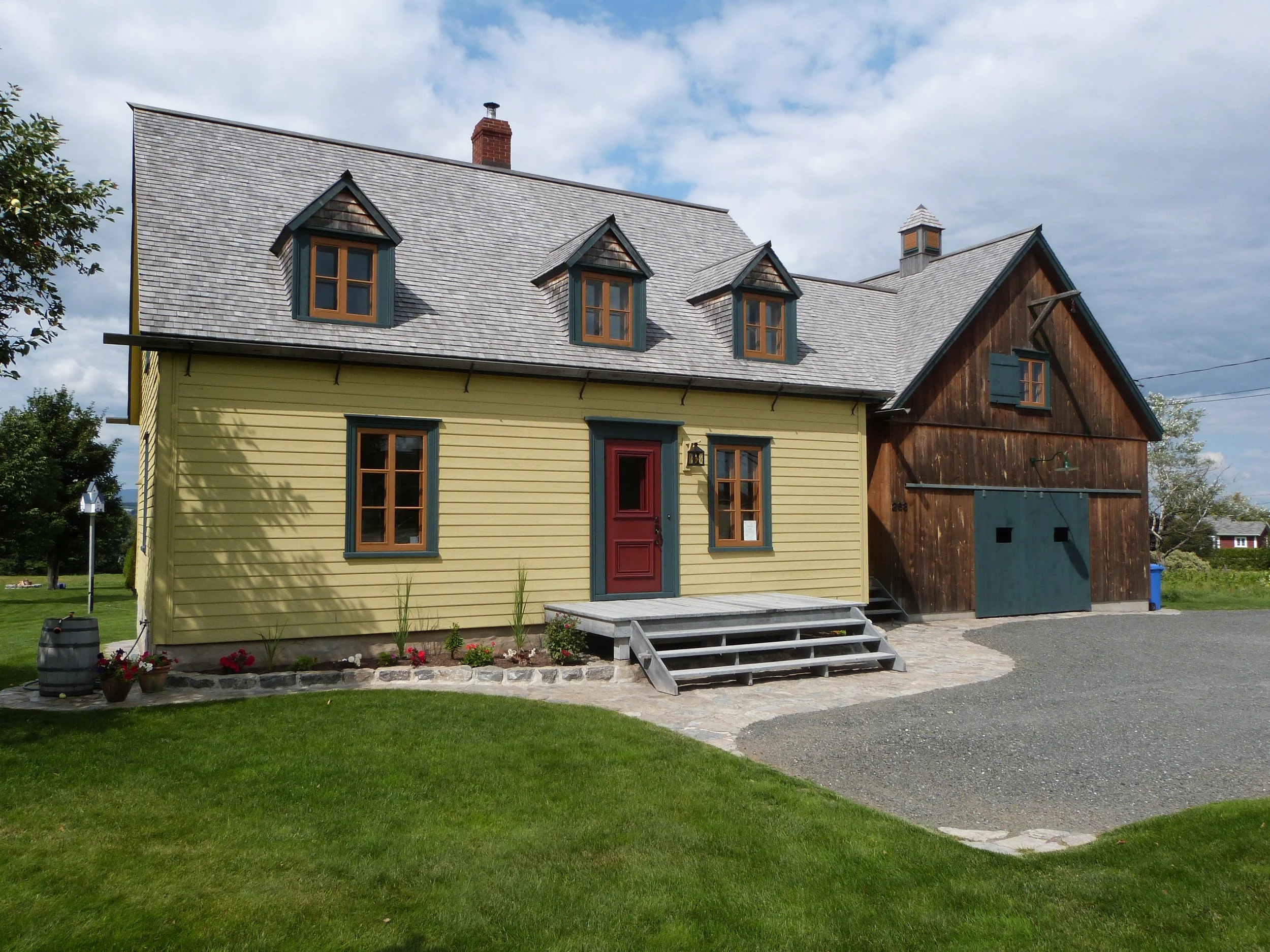 A two-story house with a yellow siding front and a dark brown wooden rear, with a gray shingle roof, set in a green yard under a partly cloudy sky.