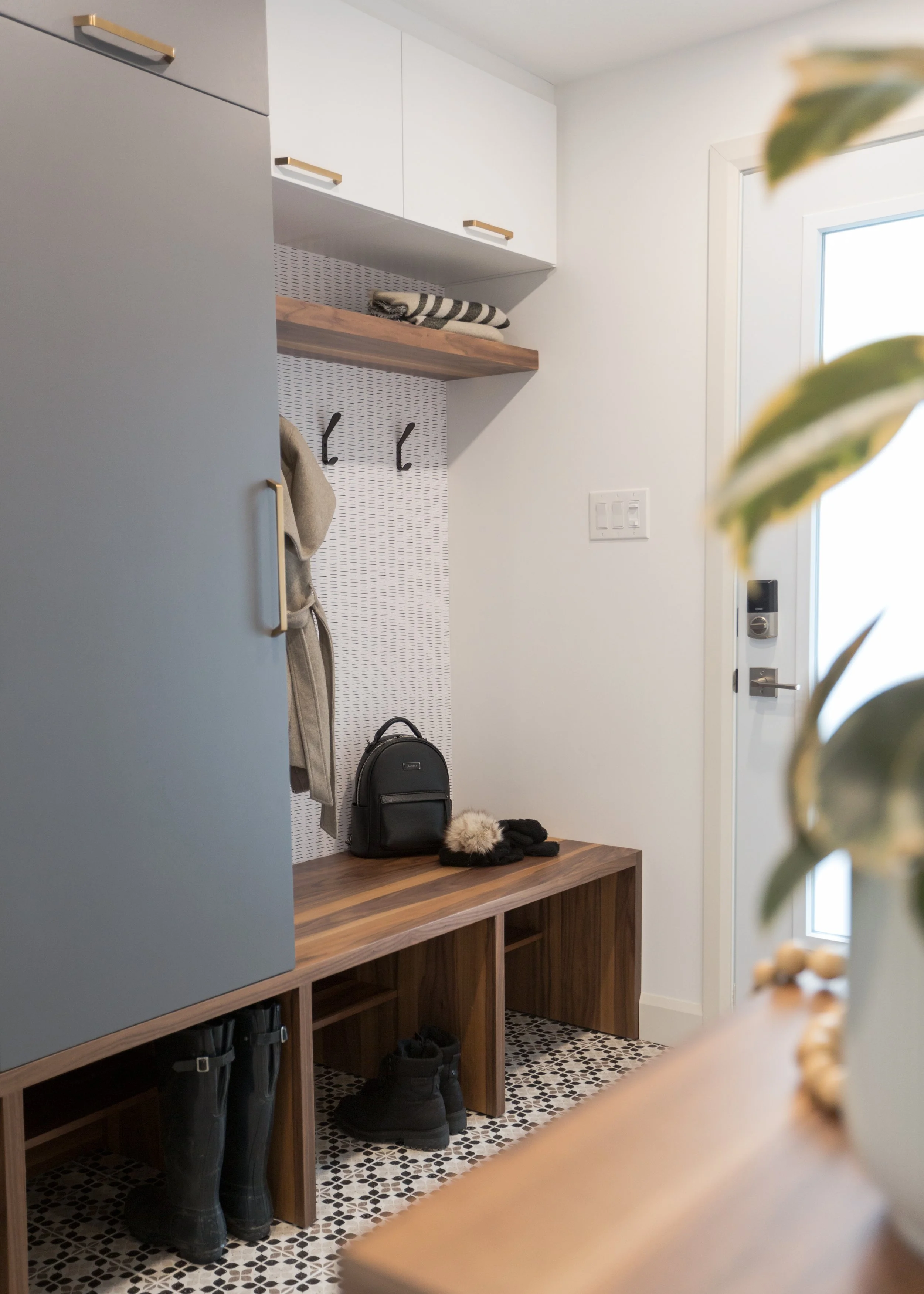 Entryway with a bench, shoe storage, hanging hooks, and a coat. A small black backpack and gloves are on the bench. A pair of black boots is underneath. The door has a keypad lock and a window.
