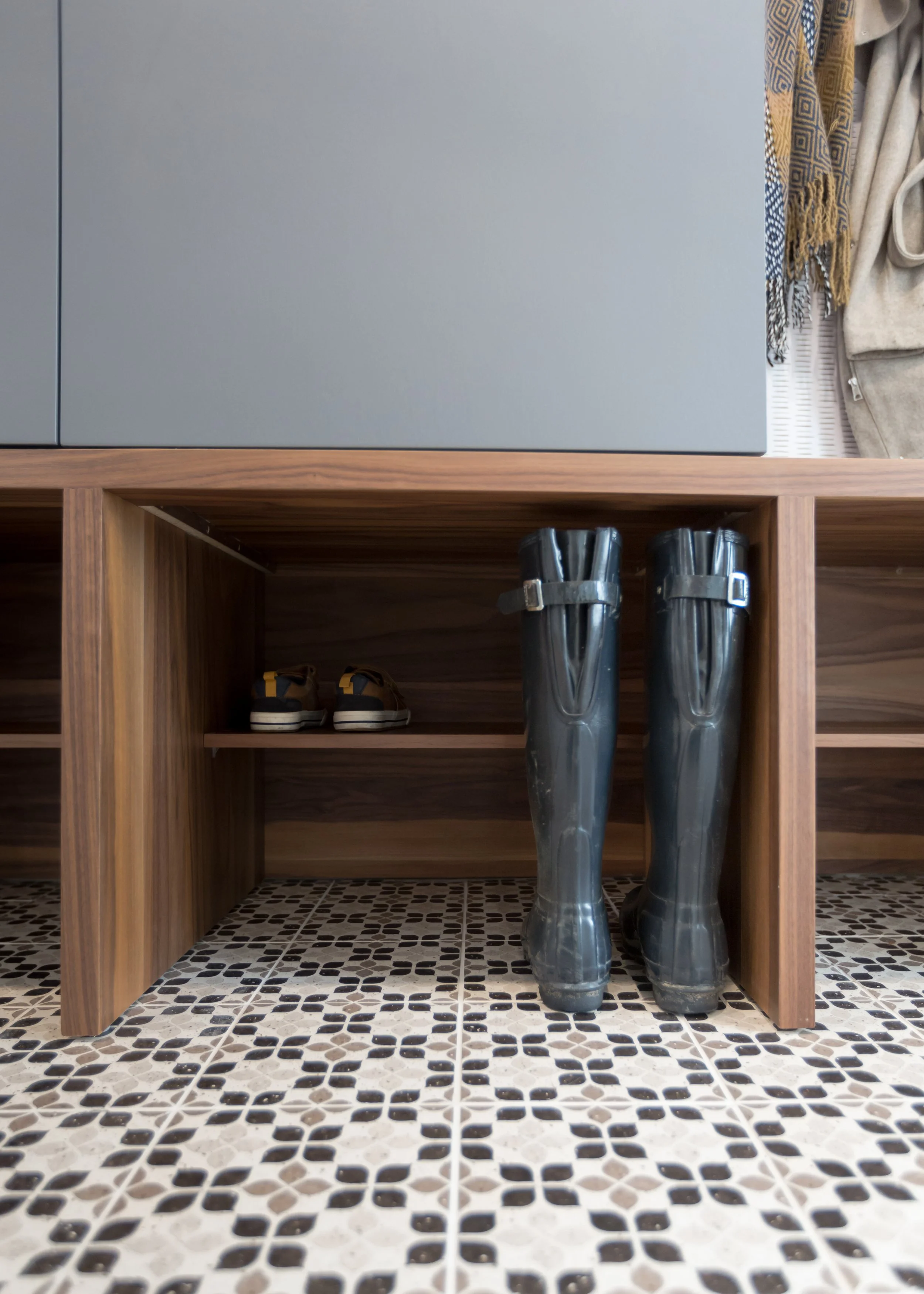 Pair of black rain boots and a pair of sneakers stored under a wooden bench in an entryway.