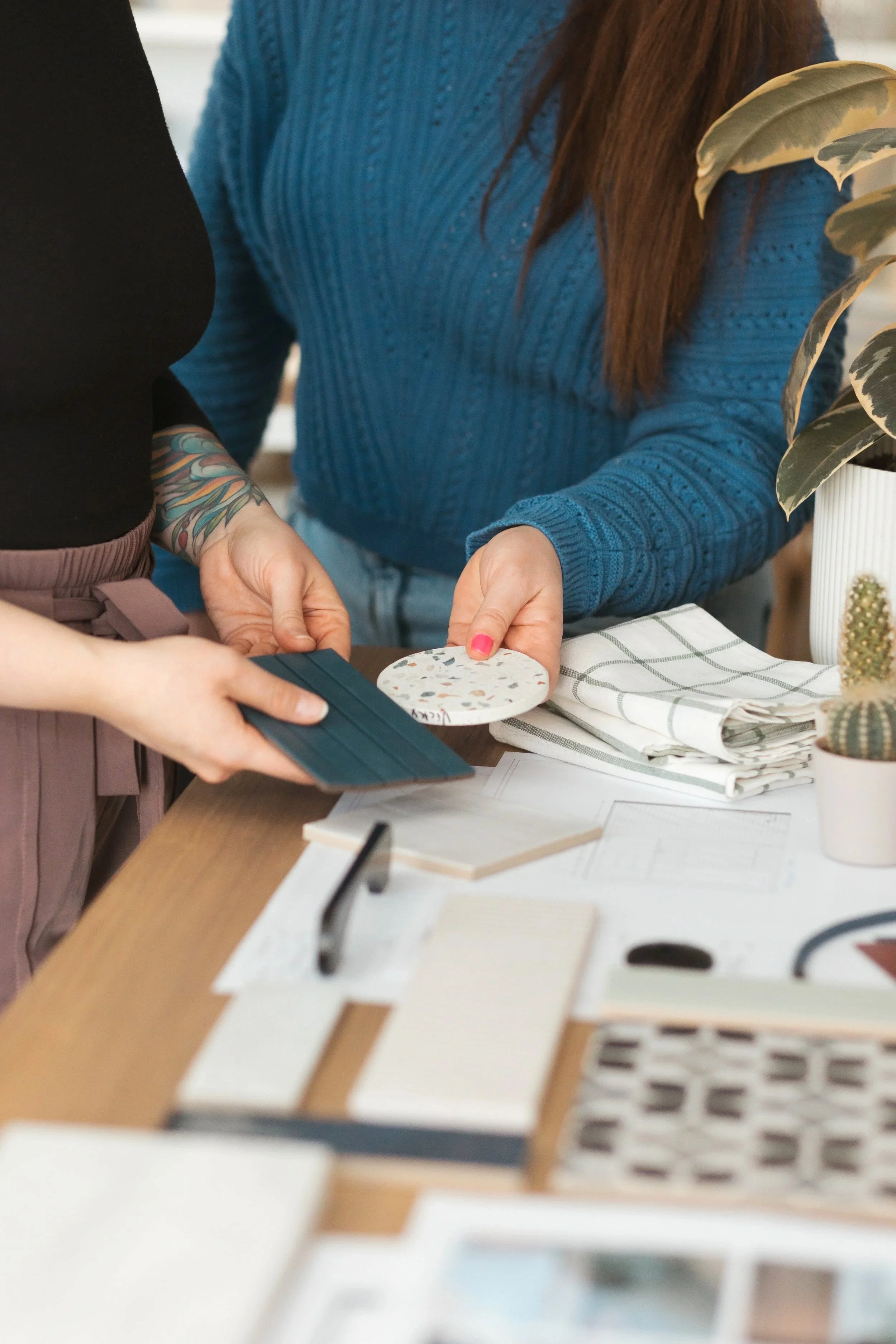 Two women are examining fabric color and pattern samples at a table with various fabric swatches, papers, and potted cacti.