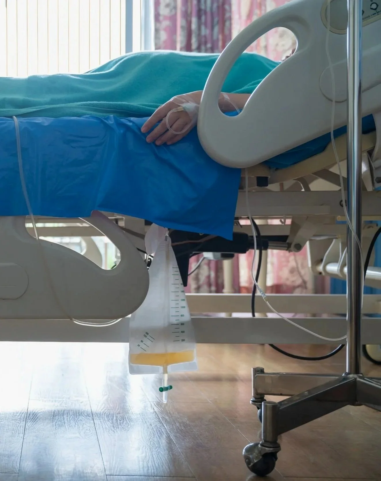 Hospital bed with a patient’s hand resting on the side, monitors, and IV fluids in a medical room with curtains.