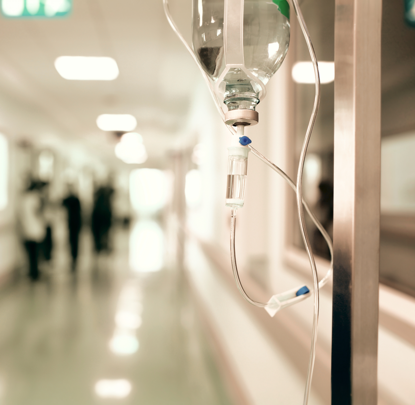 Close-up of an IV drip attached to a hospital stand in a hallway with blurred people walking in the background.