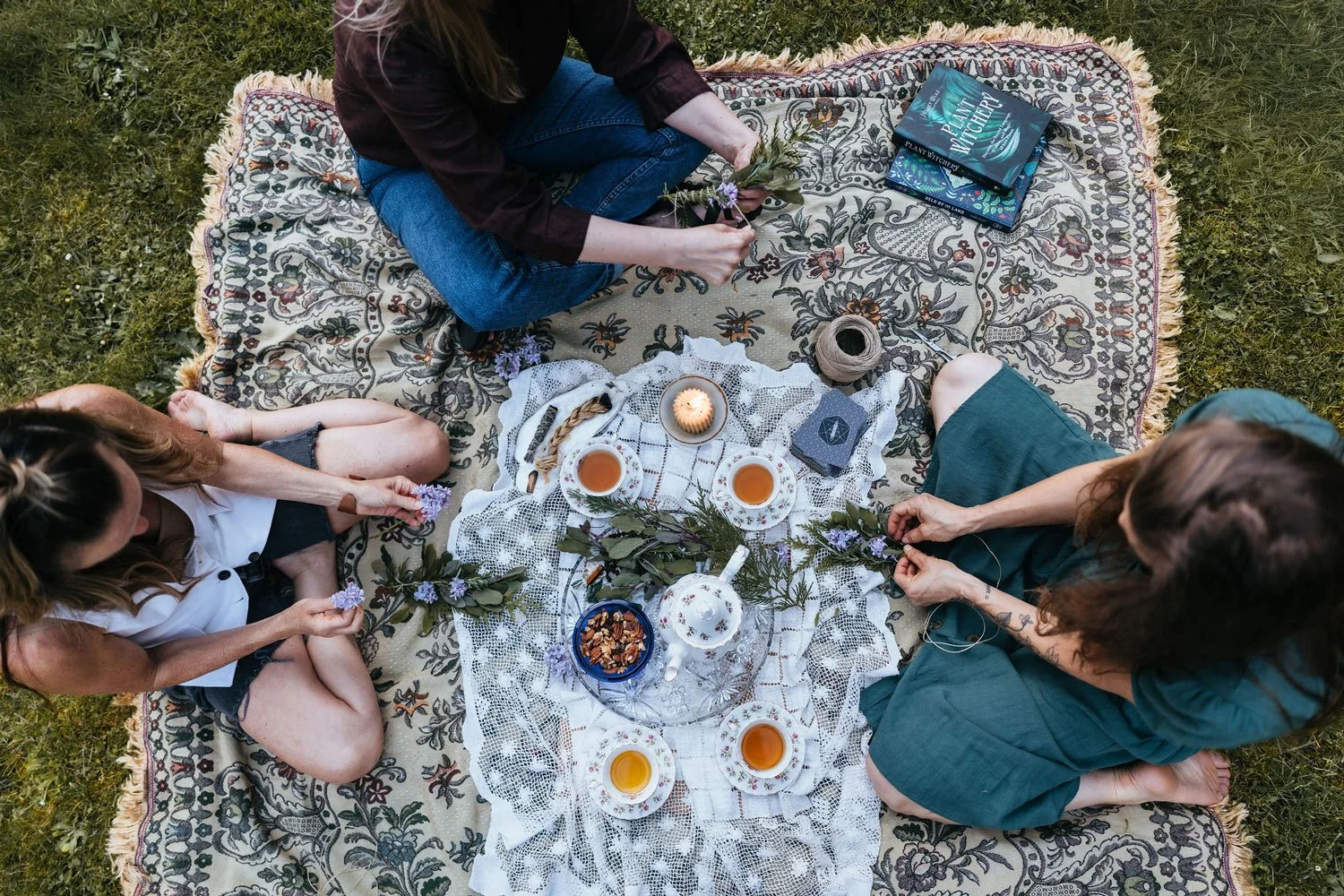 Overhead view of three women sharing herbal tea and botanicals on an ornate rug outdoors, embodying plants become art community