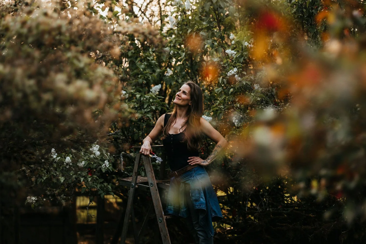 Alex, landscape designer and founder of Plants Become Art, leaning confidently on a wooden ladder surrounded by a blossoming orchard on Vancouver Island
