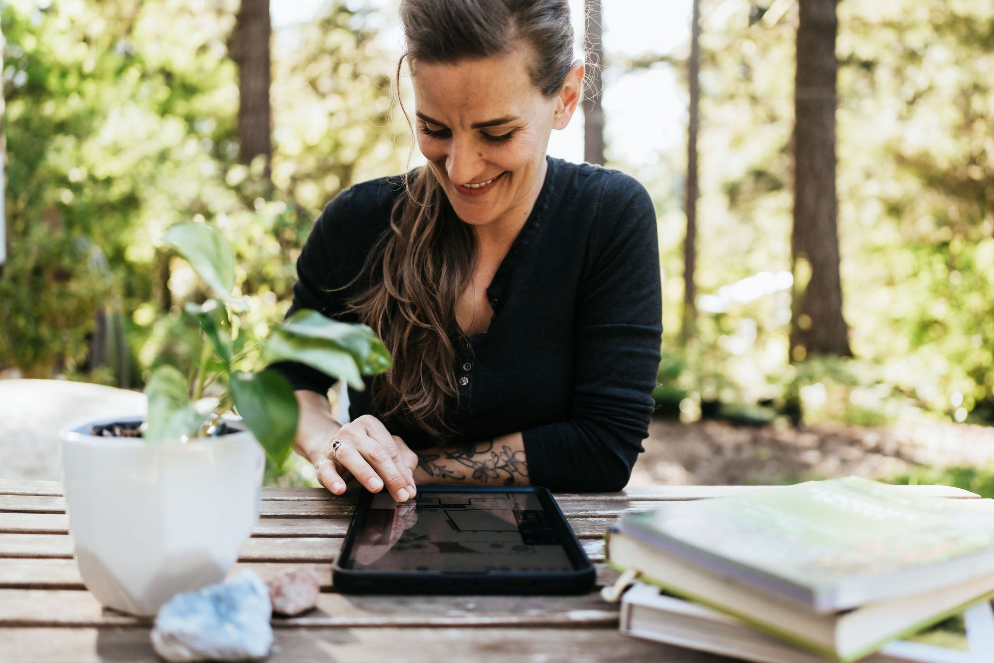 Alex smiling at her tablet on an outdoor table with a houseplant, crystal, and garden notebooks, preparing an online garden consultation
