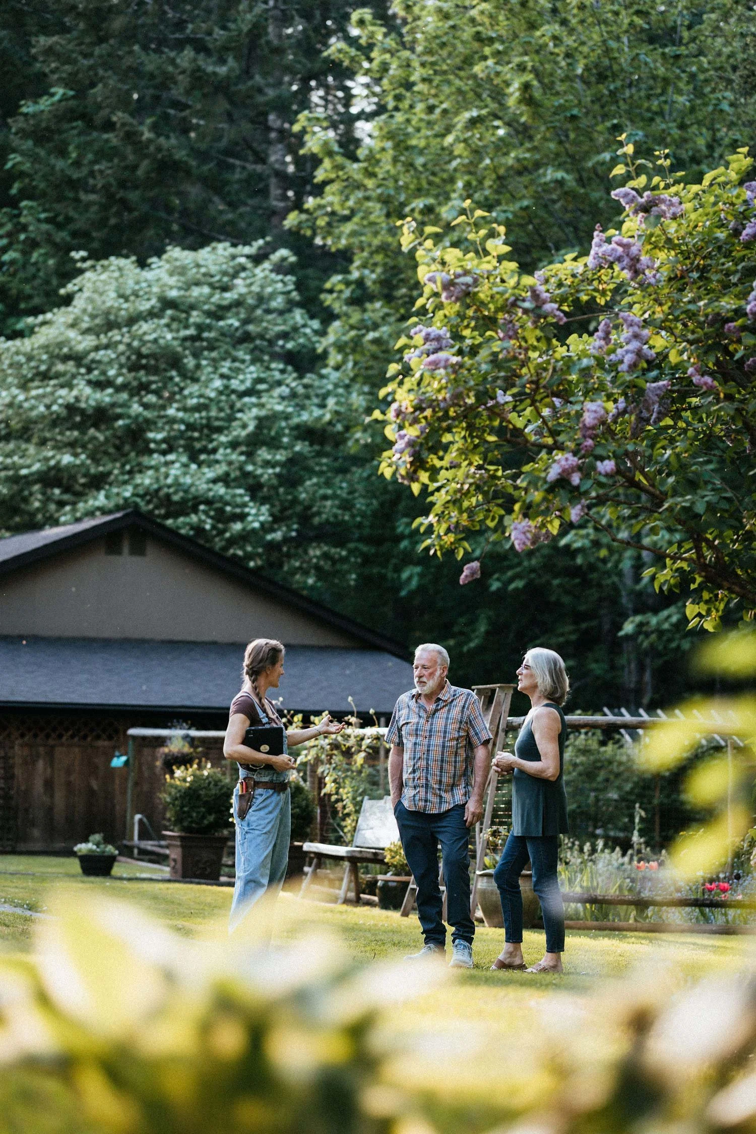 Alex consulting with homeowners in their lush garden, with lilac trees and raised beds in the background, Cowichan Valley
