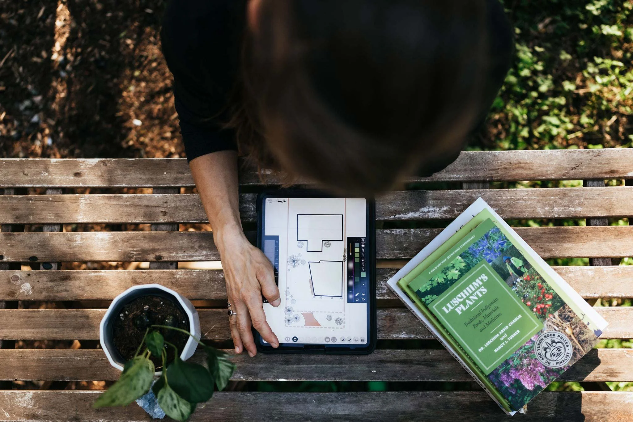 Overhead view of a custom garden design plan on a tablet beside Luschhim's Plants reference book on an outdoor bench