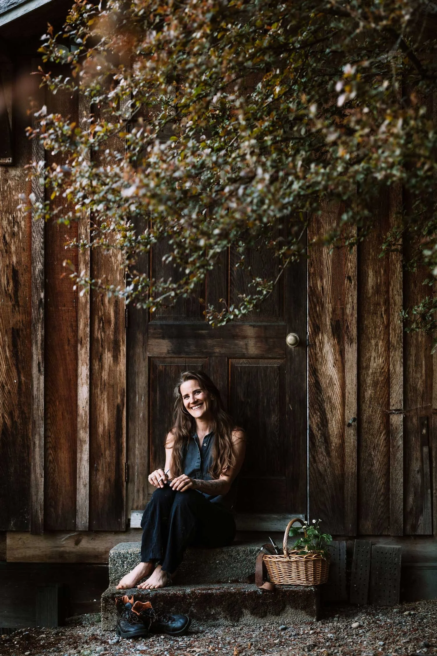 Alex, founder of Plants Become Art, sitting barefoot on rustic cabin steps with a garden basket, smiling warmly on Vancouver Island