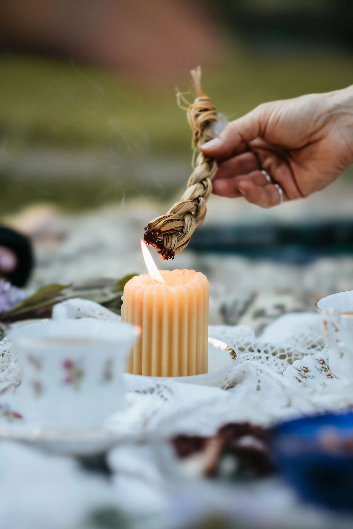 Hand lighting a braided sweetgrass bundle from a beeswax candle at an outdoor garden gathering in the Cowichan Valley