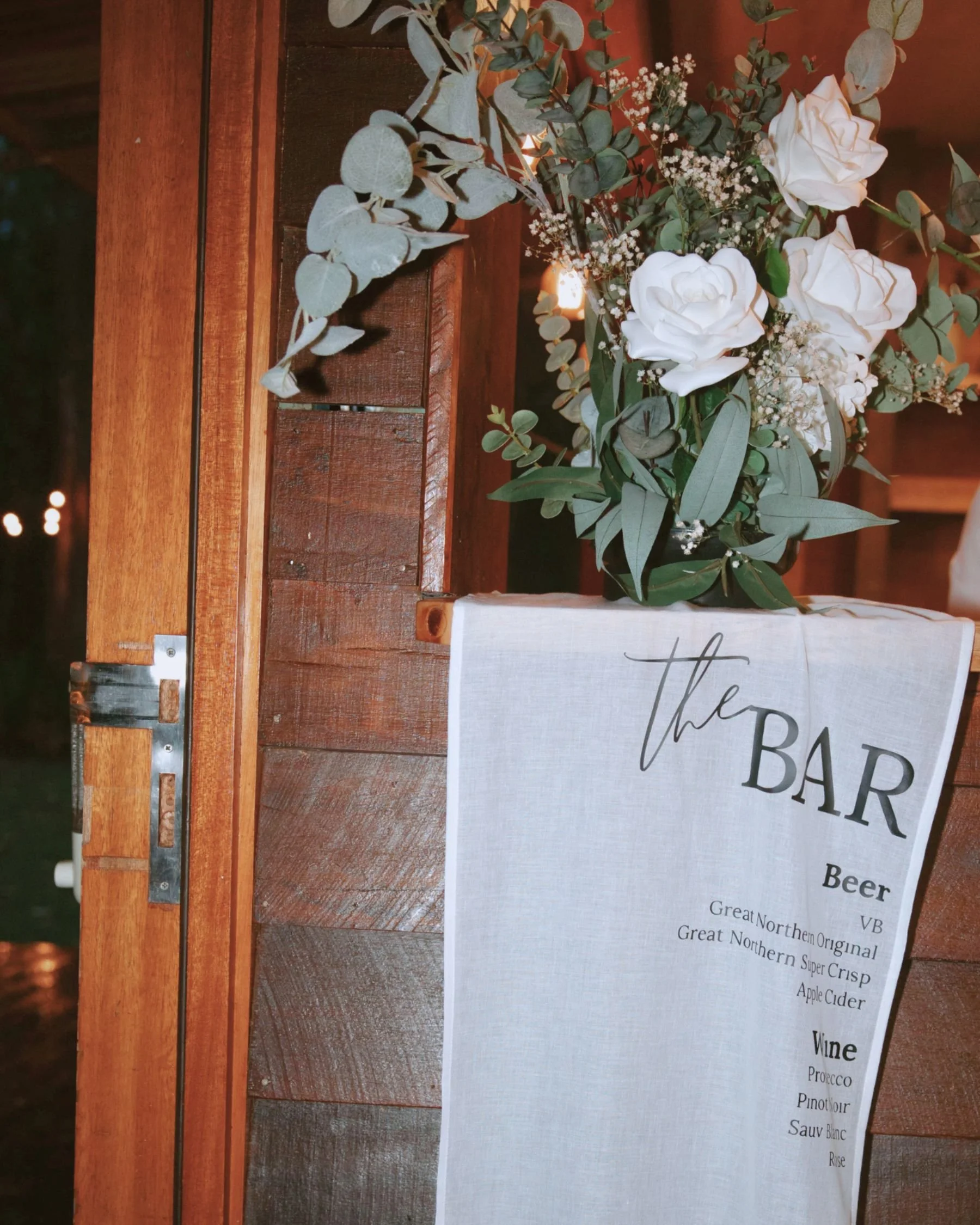 A floral arrangement of white roses, eucalyptus, and baby's breath on a wooden bar with a white cloth sign that reads 'the BAR' and lists beverages like beer and wine.