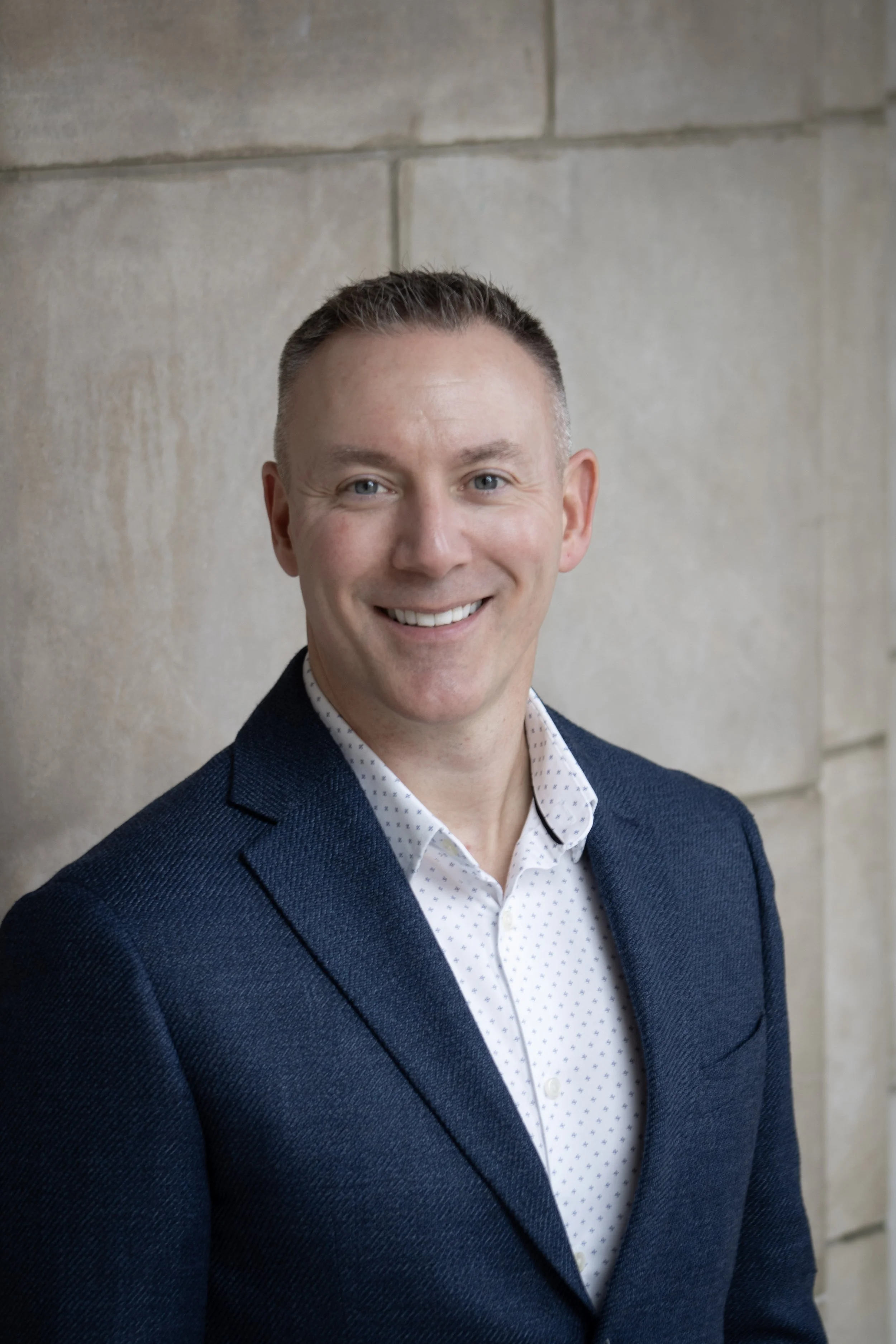 A professional headshot of a smiling man in a navy blazer and white patterned shirt, standing against a neutral stone wall background.