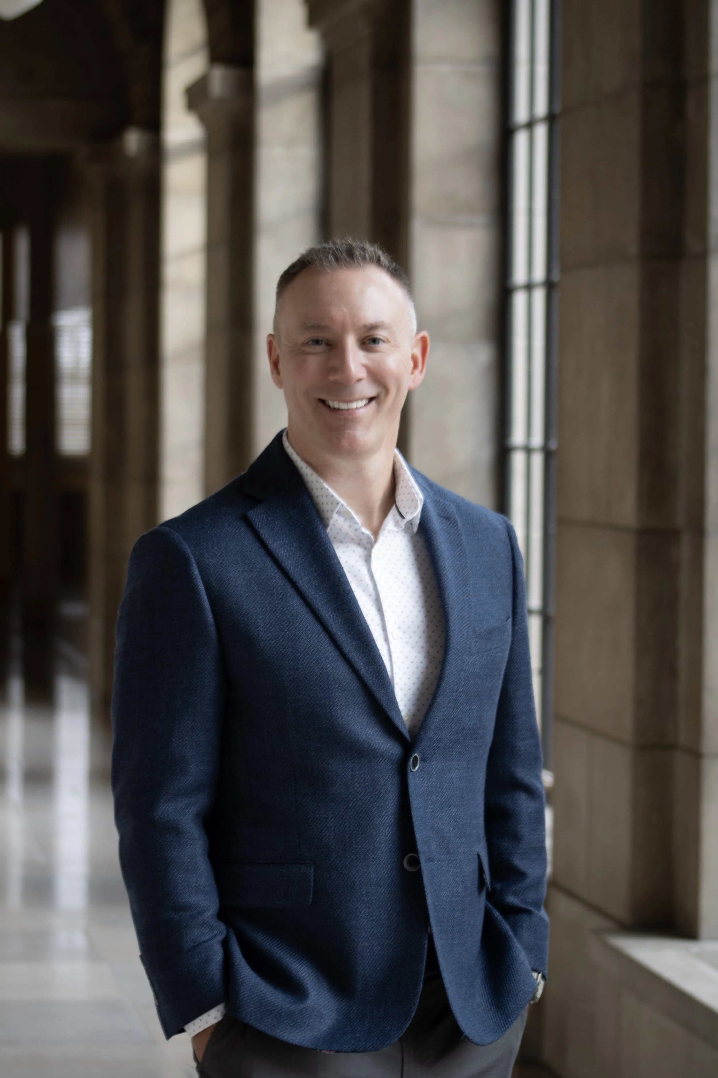 A man smiling, dressed in a navy blazer and white dress shirt, standing in a building with tall windows and stone walls. Lancaster County running for Assessor.