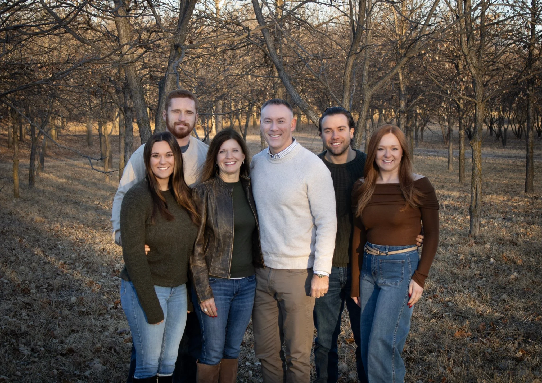 Group of six people standing outdoors in a wooded area during autumn or winter. They are smiling and dressed in casual sweaters and jeans. Doug Guess.  Guess For Assessor.