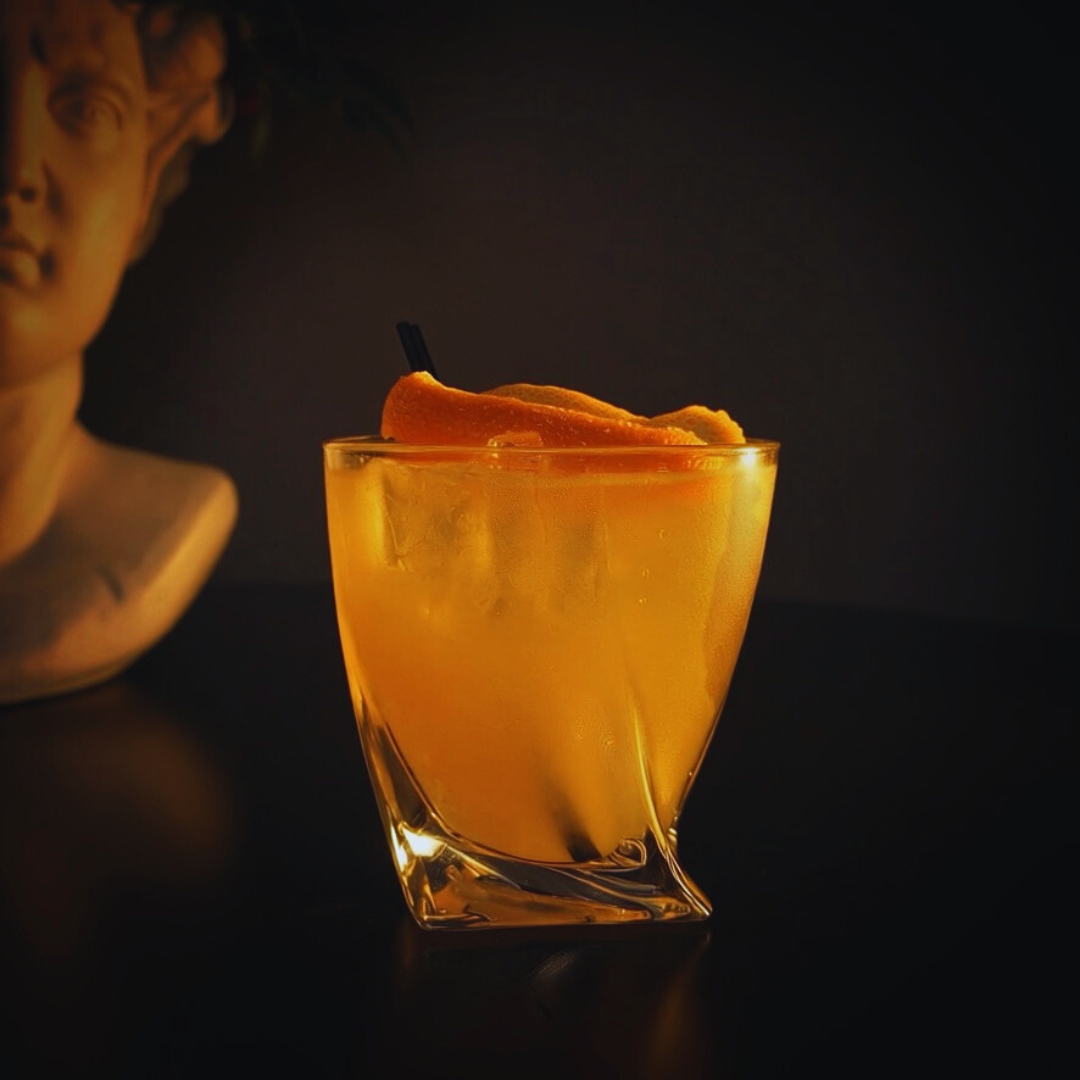 A glass of orange cocktail garnished with orange peel, with a dark background and a partially visible sculpture in the background.