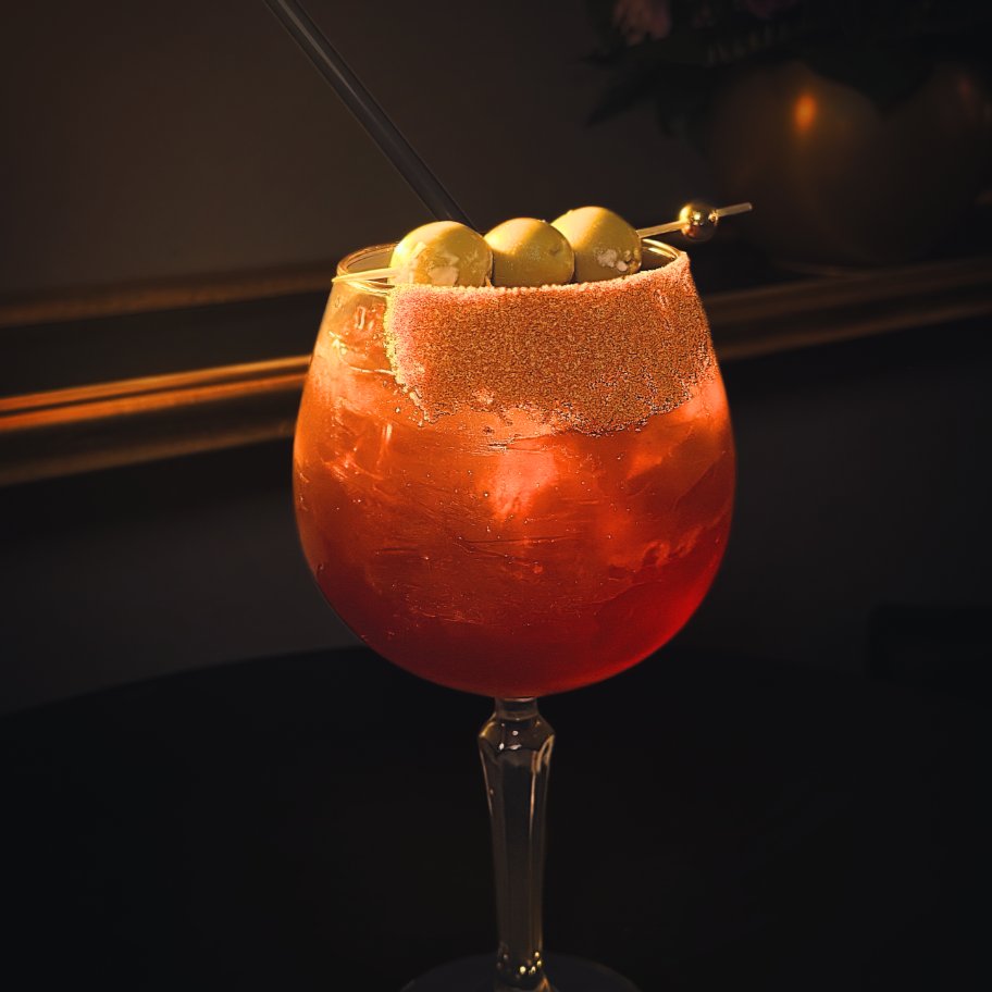 A cocktail in a large glass garnished with green olives on a skewer, with a salted rim, against a dark background.