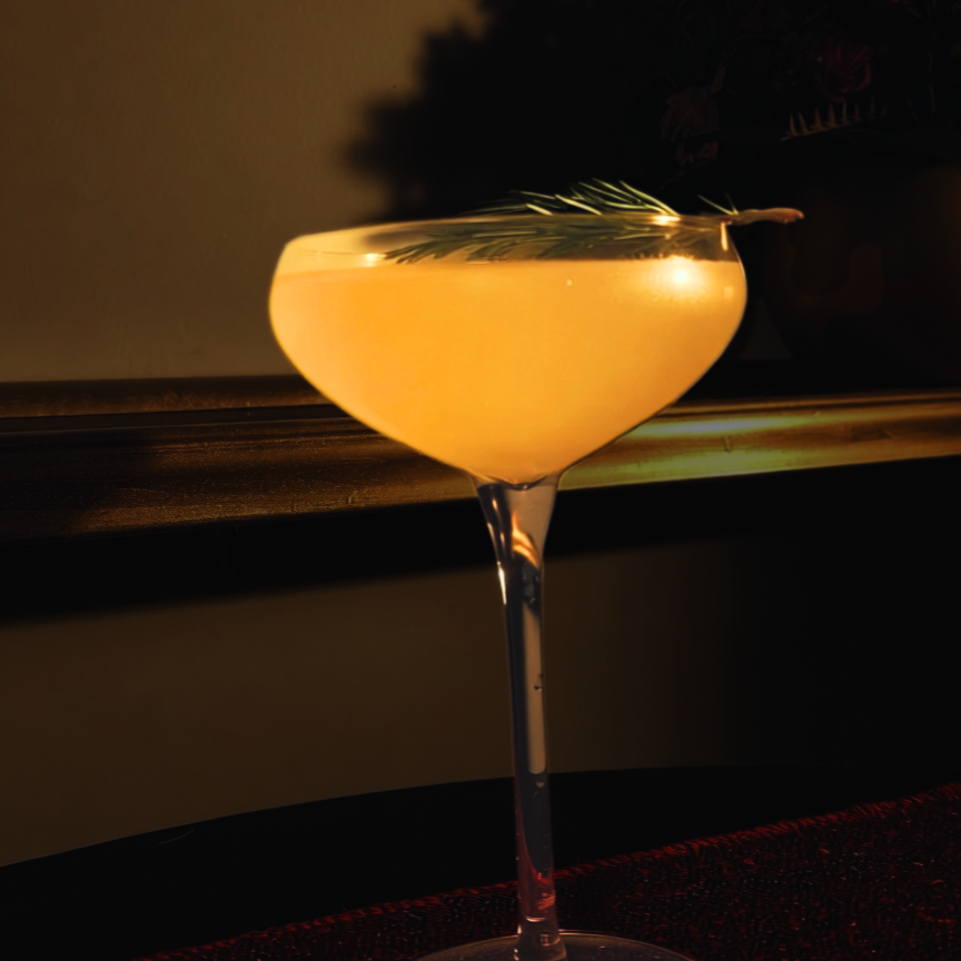 A cocktail in a coupe glass garnished with a sprig of rosemary, illuminated by warm, ambient lighting.
