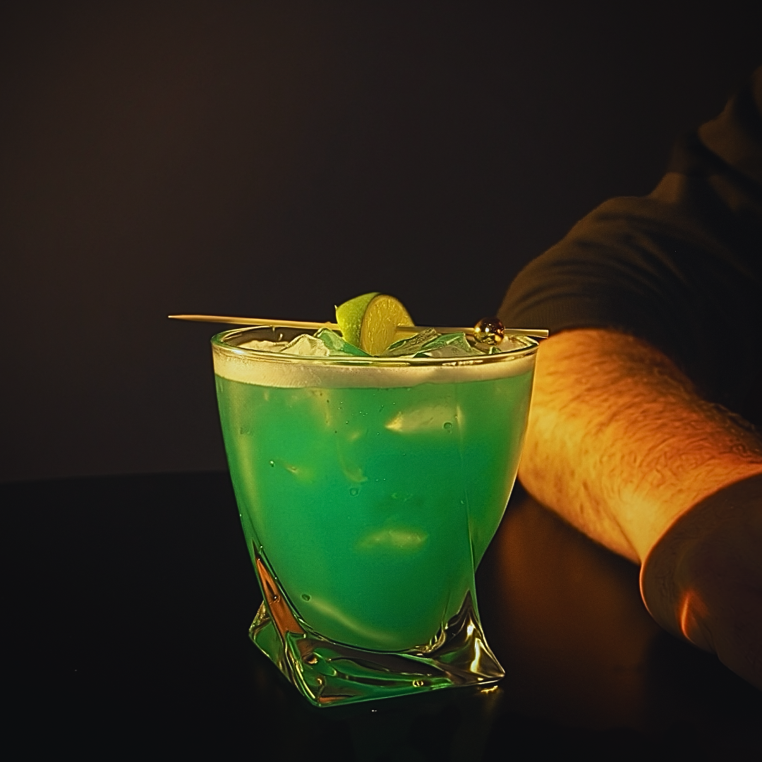A vibrant green cocktail with ice in a glass, garnished with lime and served with a cocktail pick, on a dark surface with a person’s arm resting beside it.