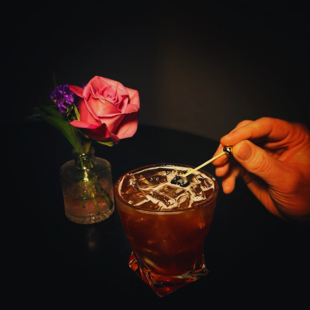 A hand is placing a cherry into a glass of iced coffee or a similar dark beverage on a black surface, with a small vase holding a pink rose and purple flowers in the background.