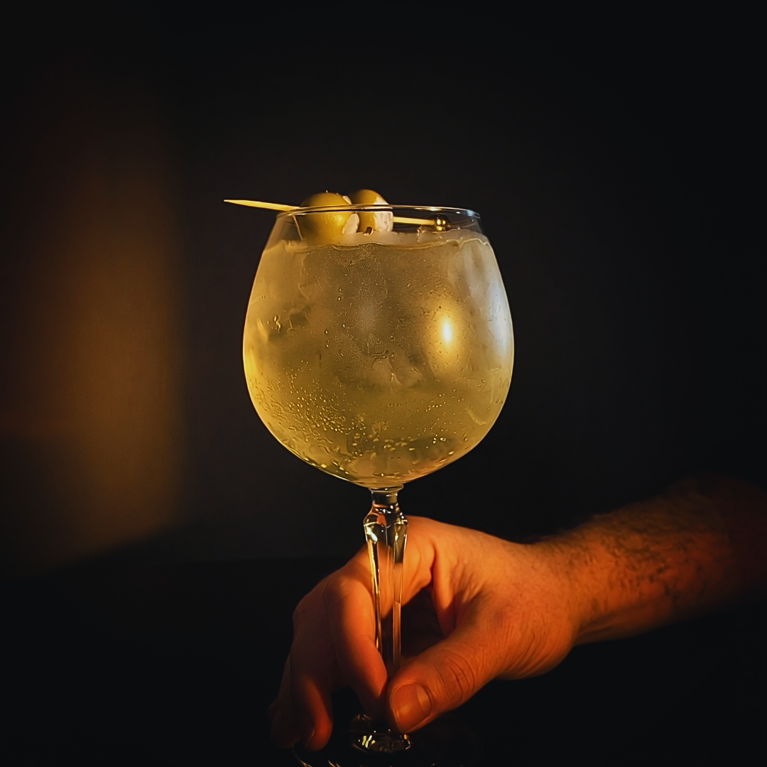 A hand holding a wine glass filled with a clear, bubbly beverage garnished with green olives on a cocktail pick, set against a dark background.