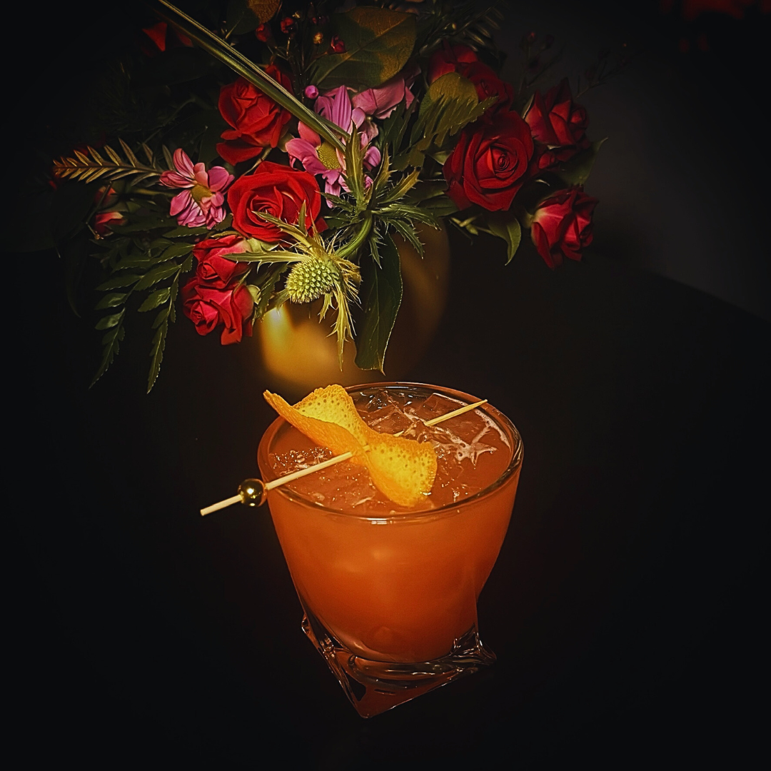 A vibrant pinkish-orange cocktail with ice cubes, garnished with an orange peel twist on a skewer. In the background, a bouquet of red roses, purple flowers, and green foliage in a yellow vase, set against a dark surface.