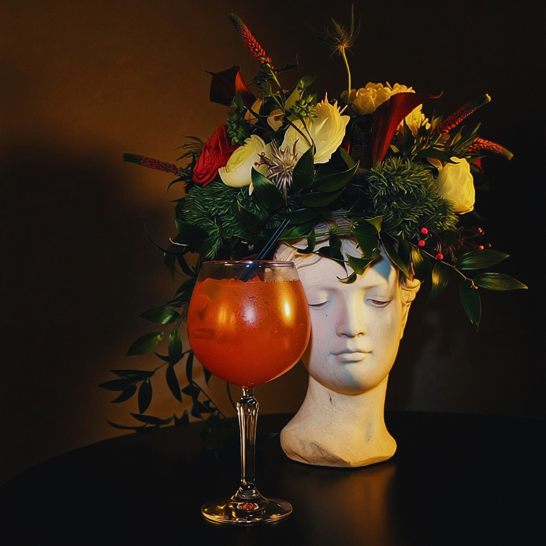 A ceramic head-shaped vase filled with yellow, red, and green flowers, with a cocktail glass containing an orange drink and a black straw placed in front.