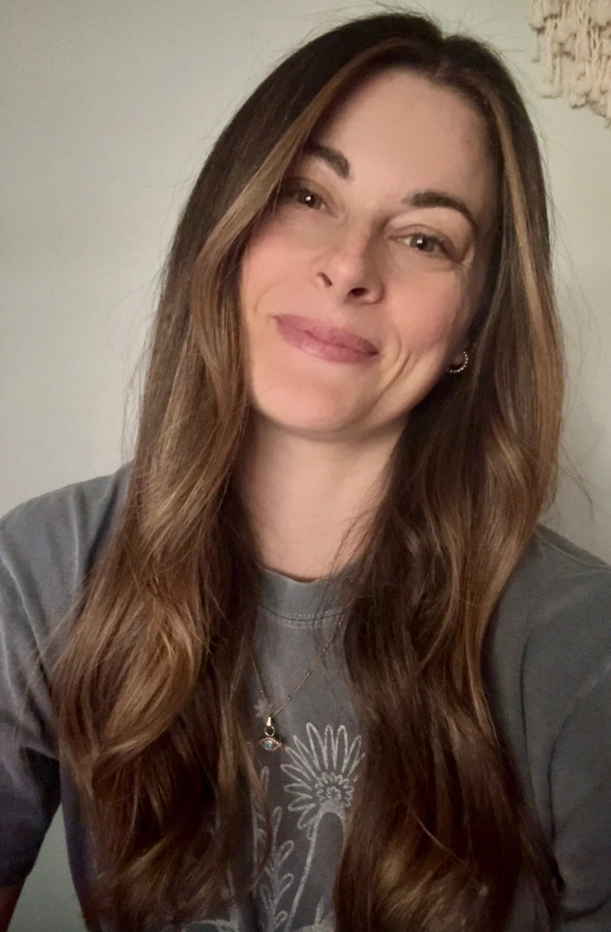 A woman with long wavy brown hair smiling, wearing a gray t-shirt and a necklace, in front of a light-colored wall with some damaged paint.