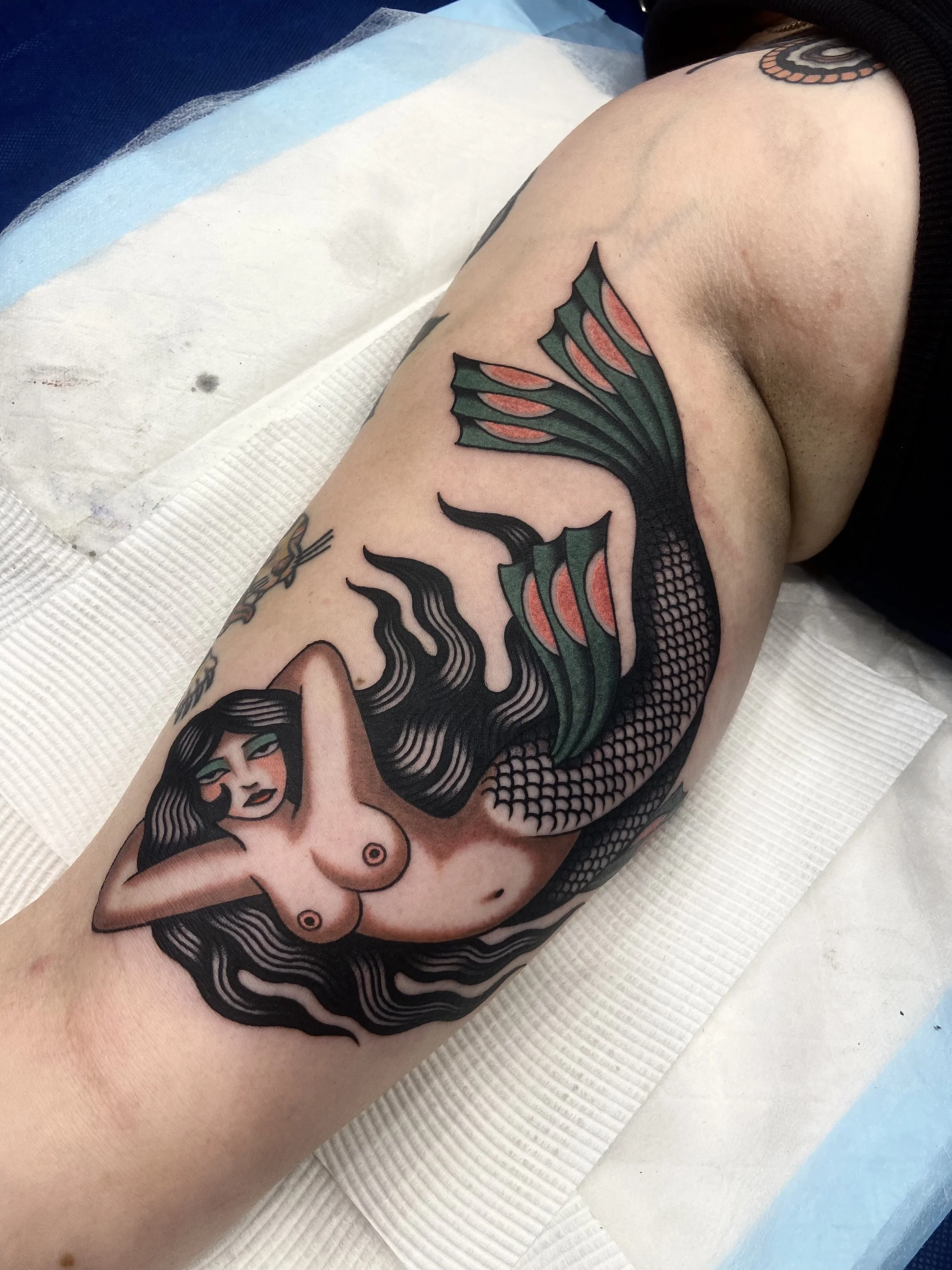 Tattoo of a mermaid with long black hair, green eyes, and a fish tail on a person's arm.