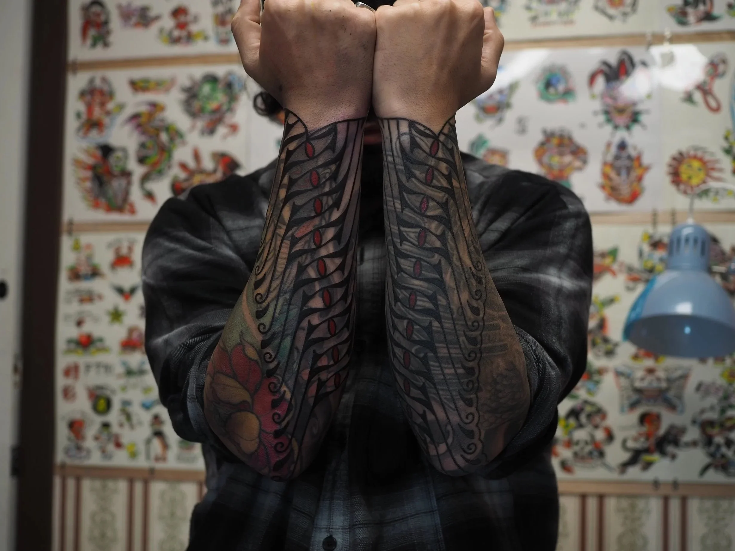 Person with tattoos on arms, covering face with fists, standing in tattoo studio with tattoo flash sheets on wall.