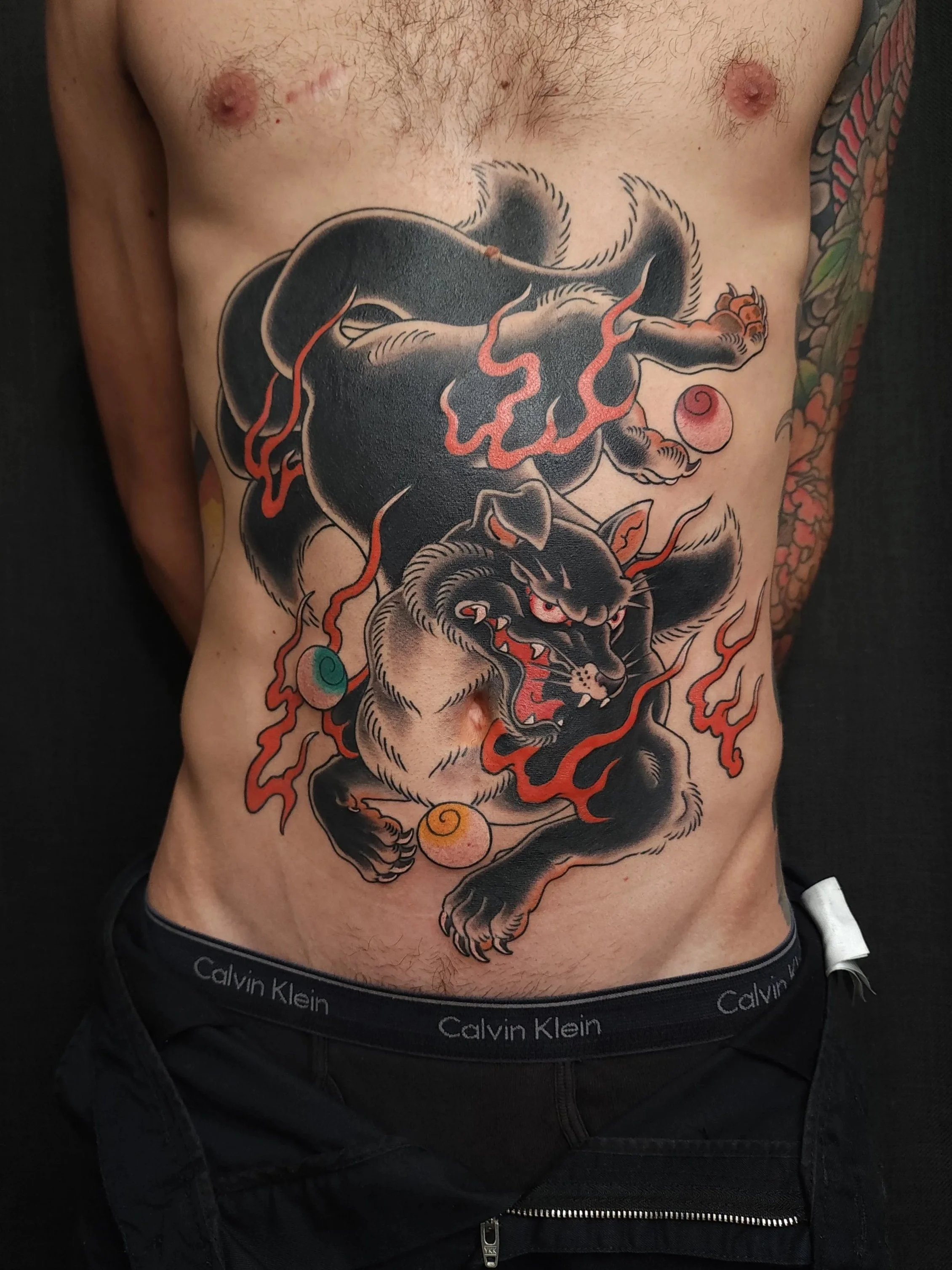 Tattoo of a fierce, traditional-style black and gray fox with red eyes, snarling and surrounded by flames, on a man's torso. The man is shirtless, revealing chest hair and tattoos on his side, and is wearing black Calvin Klein underwear.