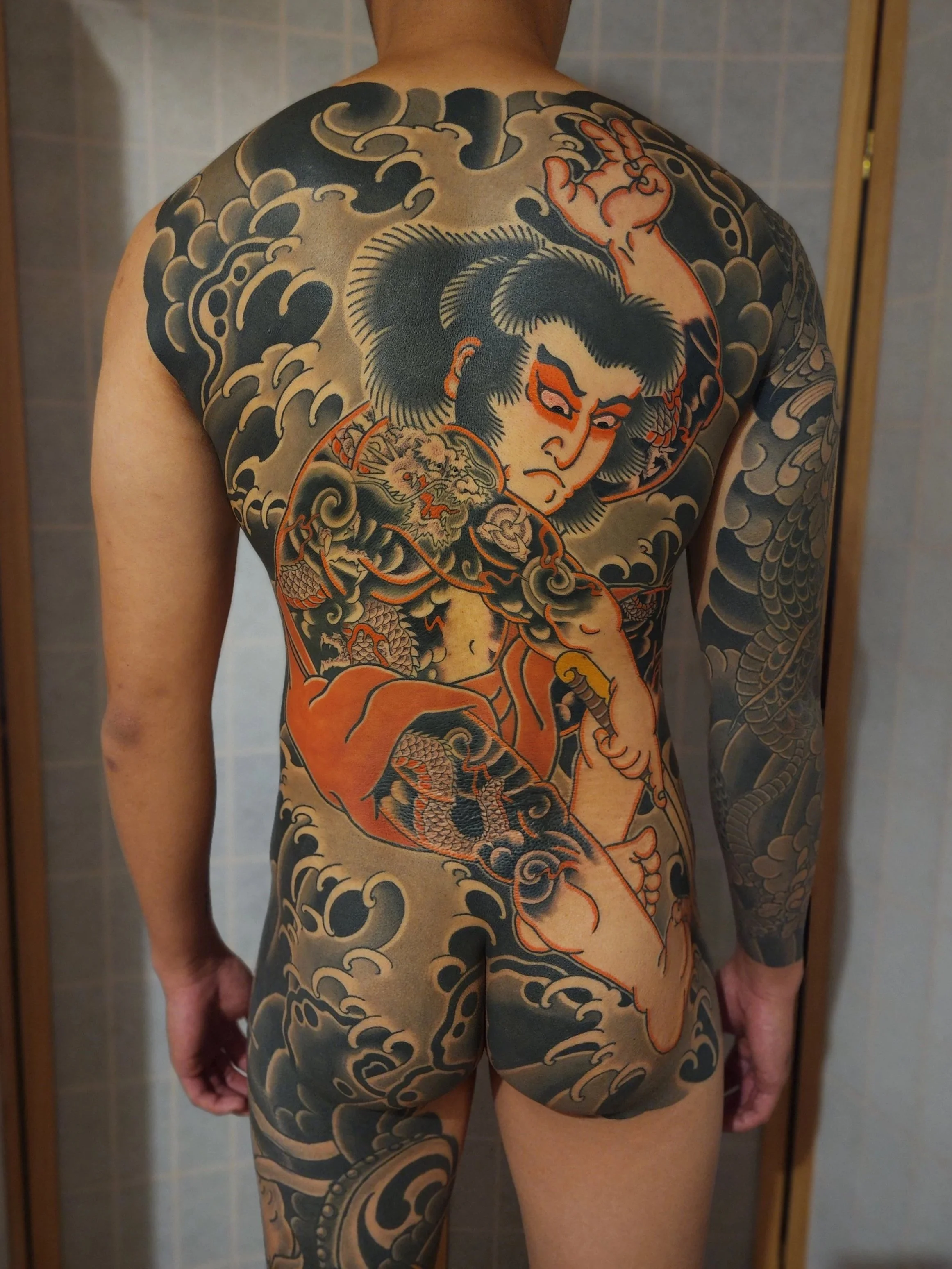 Full-back tattoo of a traditional Japanese style, depicting a fierce face of a warrior with long hair, surrounded by waves, clouds, and dragons, with detailed line work and shading.