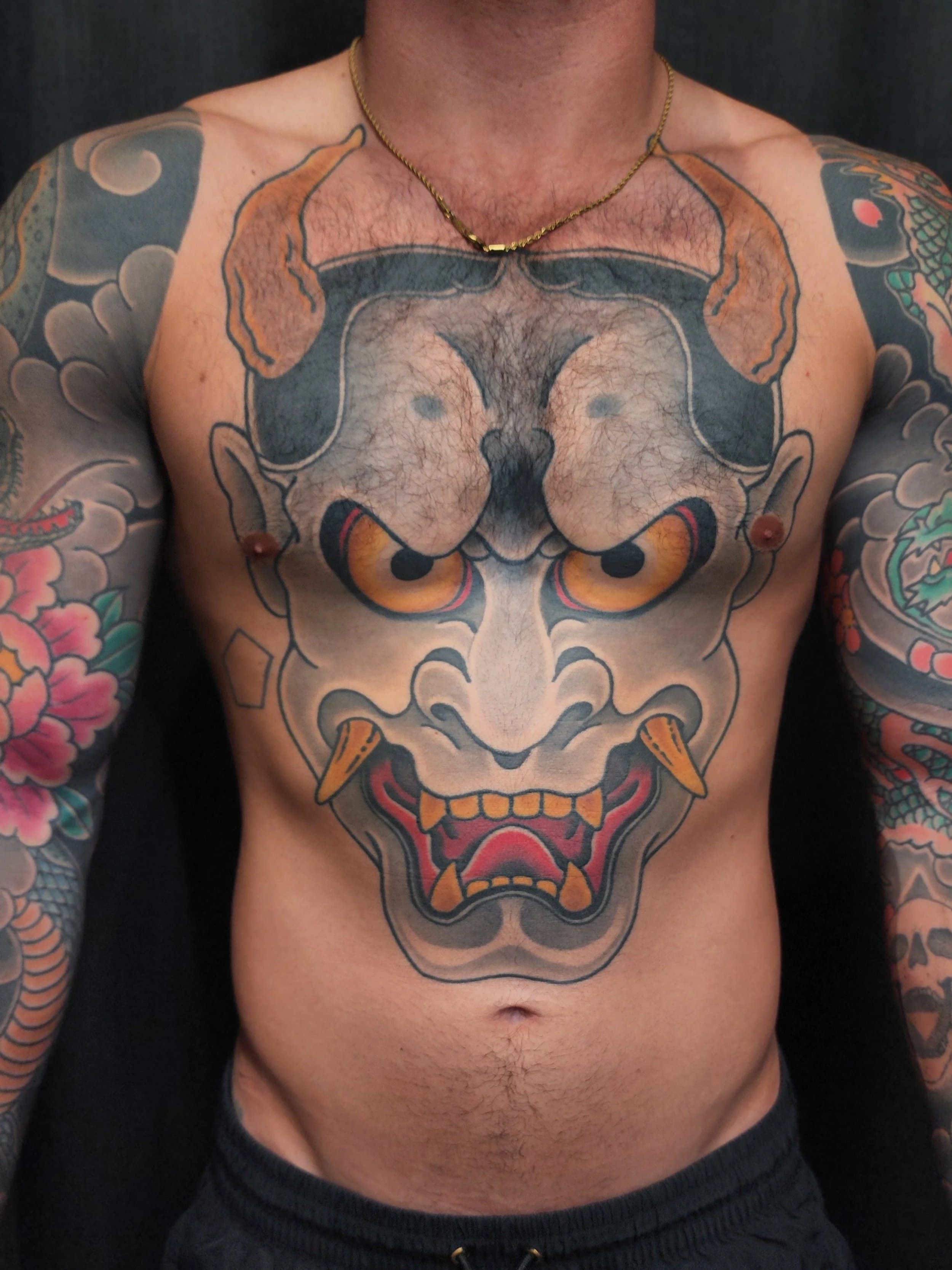 A tattoo of a fierce Hannya mask with exaggerated features on a man's chest, surrounded by colorful tattoos on his arms.