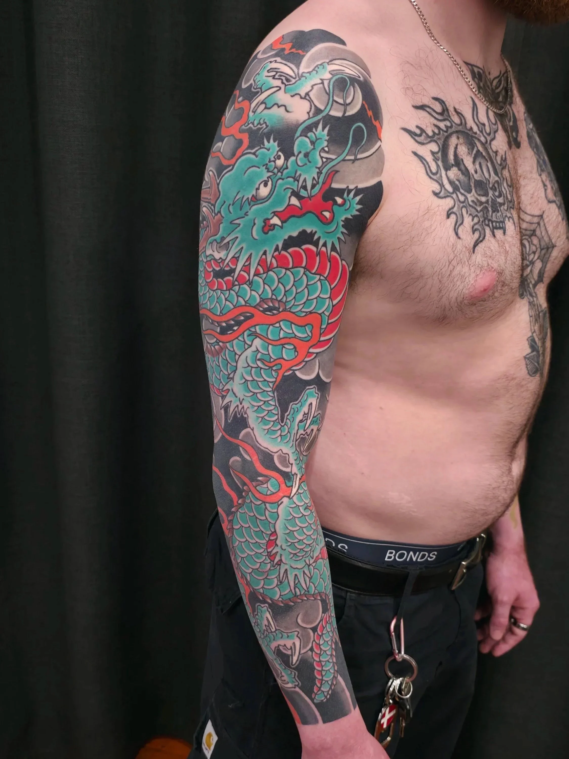A person with an elaborate Japanese-style tattoo of a dragon on their left arm, featuring blue, red, and black ink, with the dragon's head near the shoulder and its body extending down to the wrist.