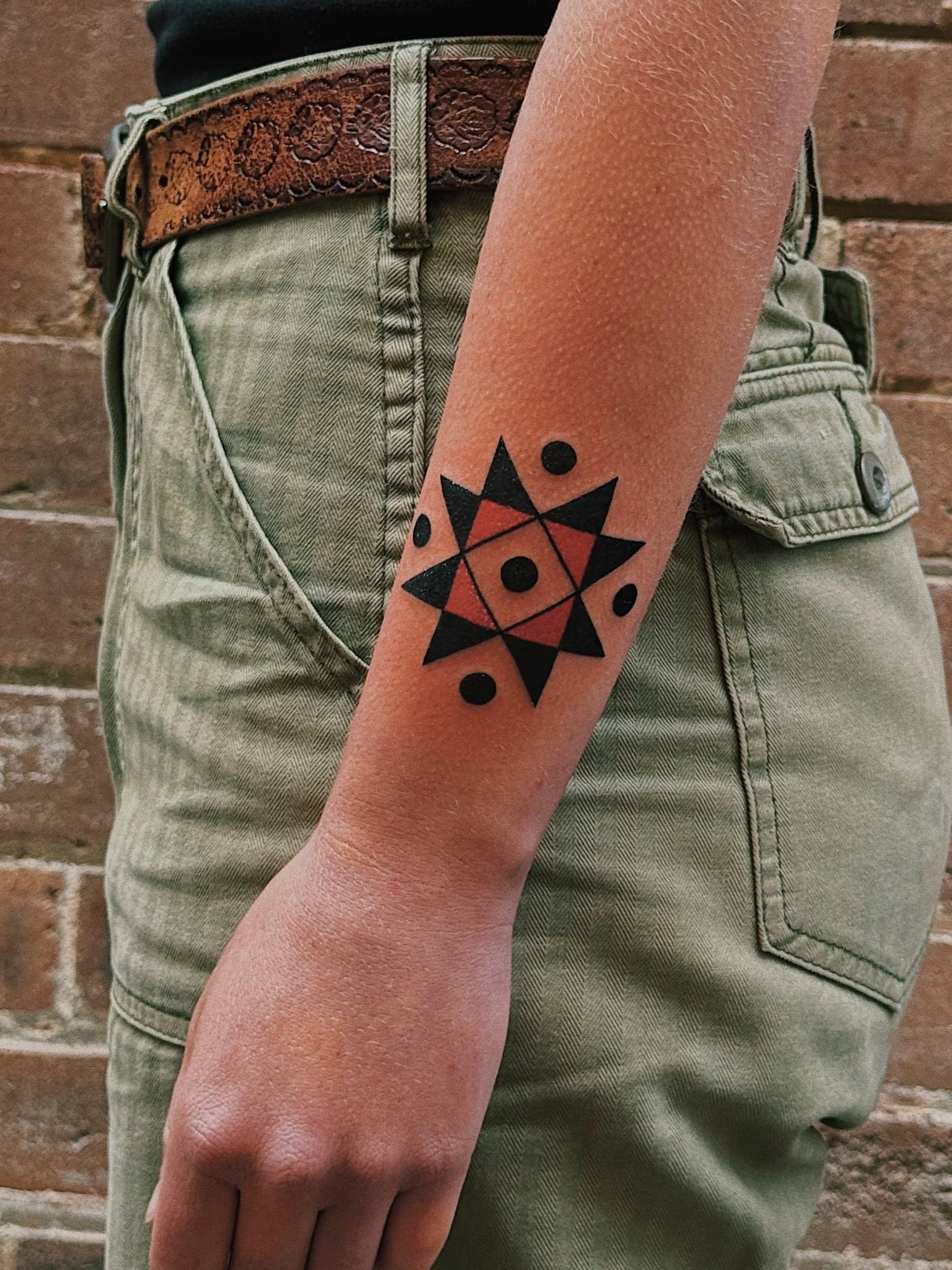 A person's forearm displays a geometric tattoo with black and red shapes, including dots and triangles, against a brick wall background.
