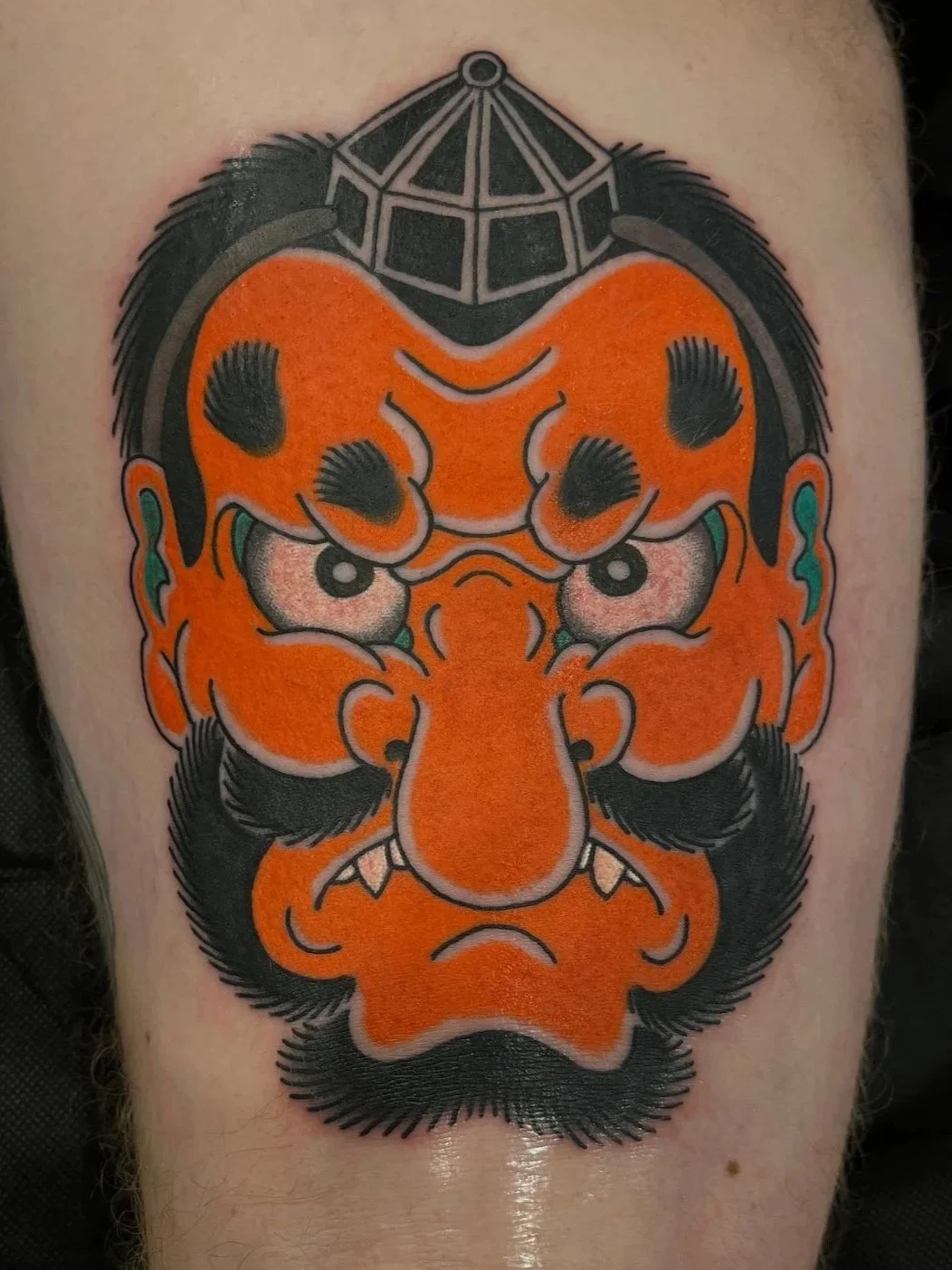 Traditional Japanese-style tattoo of a fierce, stylized tengu with bright orange skin, black hair, eyebrows, mustache, and beard, and a forehead ornament resembling a crown or helmet.