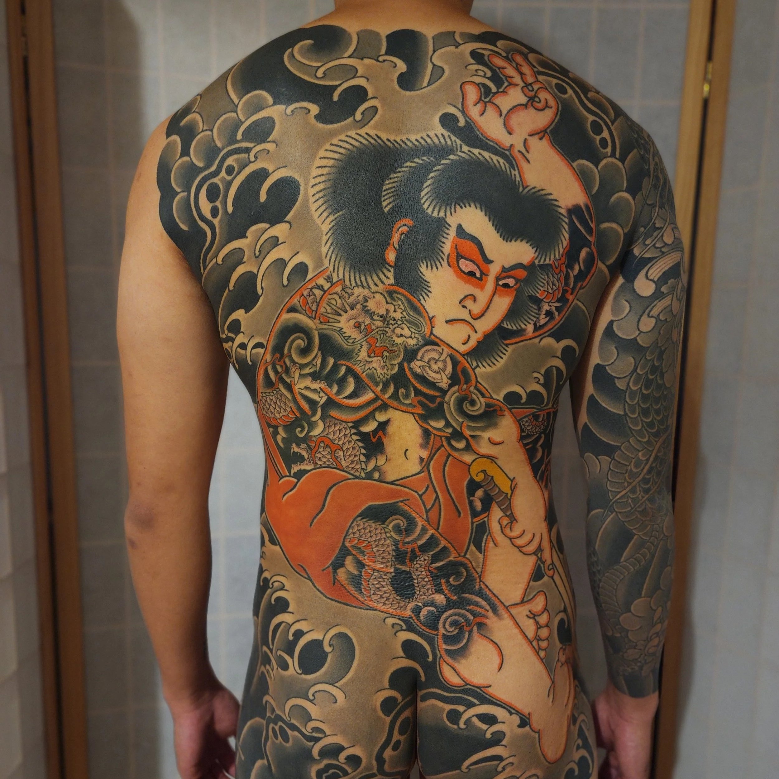 Back of a person with a large traditional Japanese style tattoo featuring a fierce warrior, a dragon, and swirling clouds.