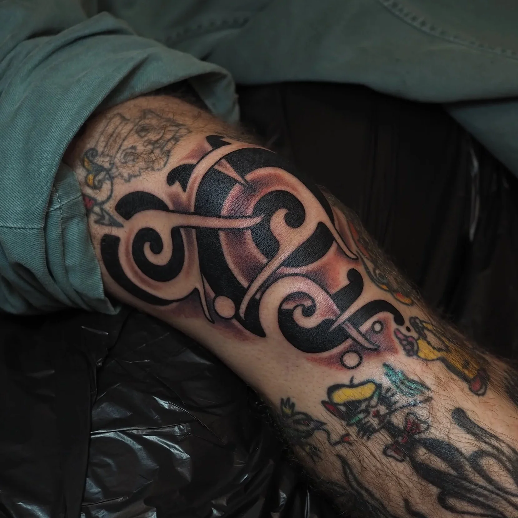 Close-up of a person's arm with black tattoos, including a large black and grey tribal-style design and smaller cartoon-like characters.