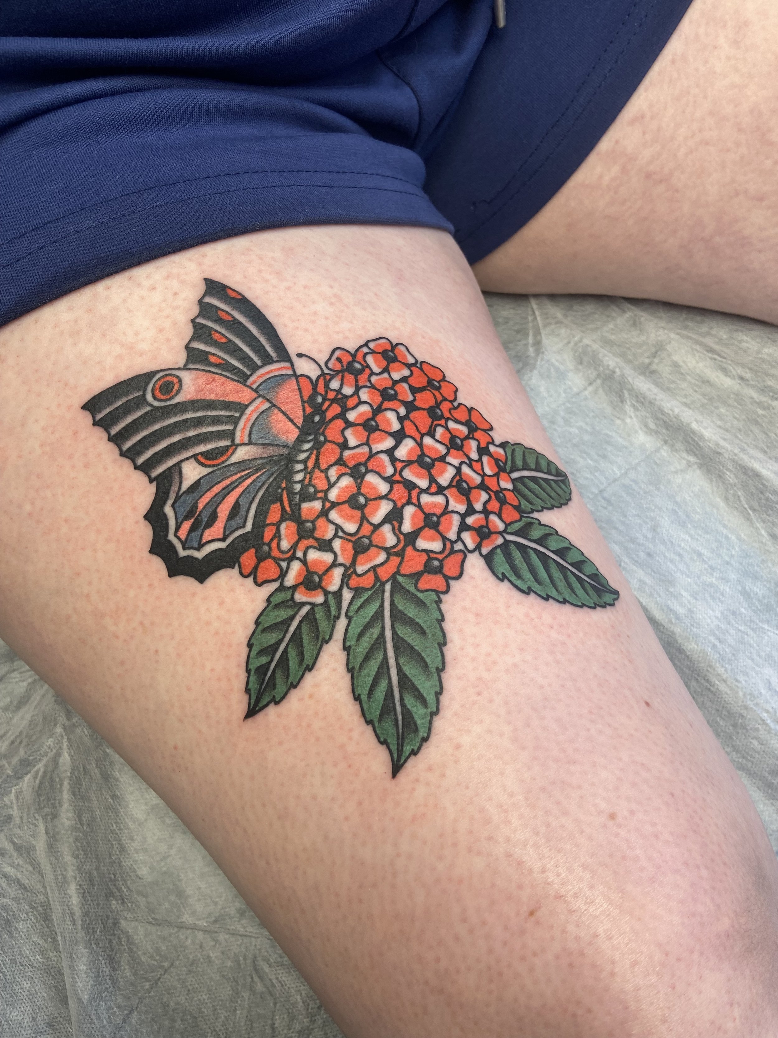 Tattoo of a butterfly perched on a cluster of orange and pink flowers with green leaves on a person's thigh.