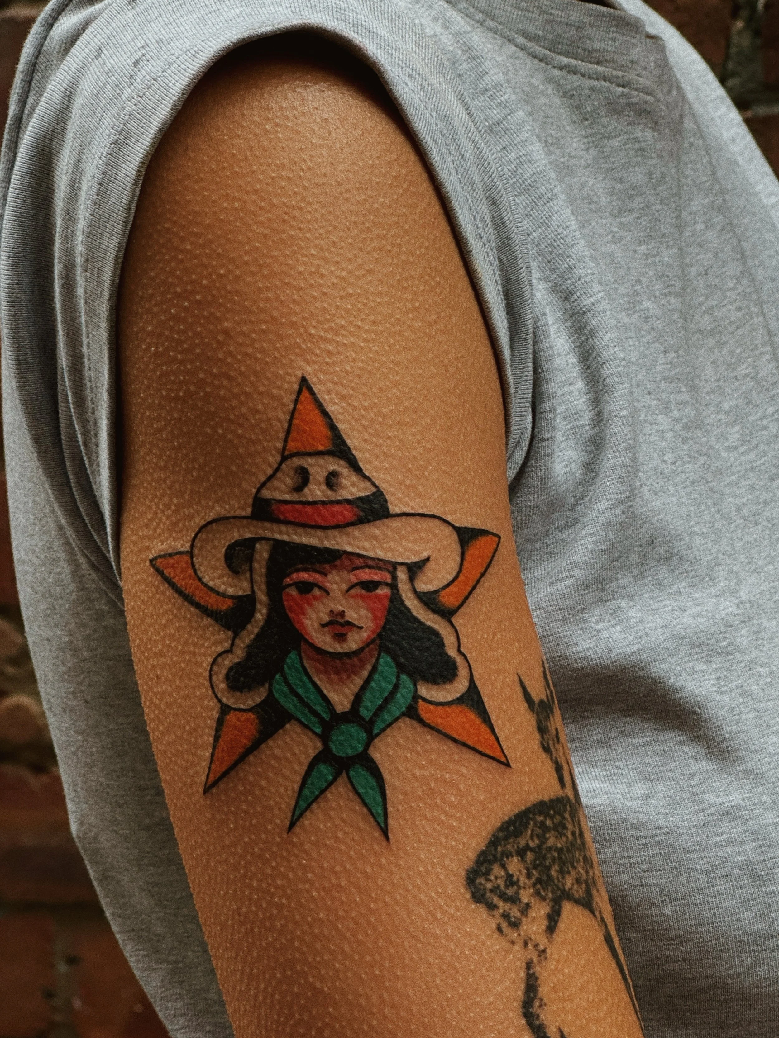 Tattoo of a woman wearing a cowboy hat, with a green scarf, placed on an orange star on the upper arm.