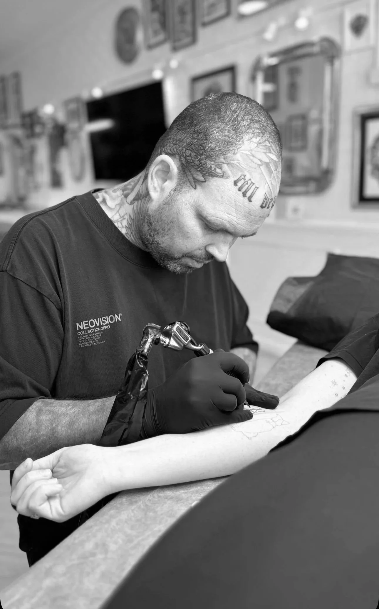 Tattoo artist in black gloves tattooing a person's forearm in a tattoo parlor, with framed pictures on the wall in black and white.