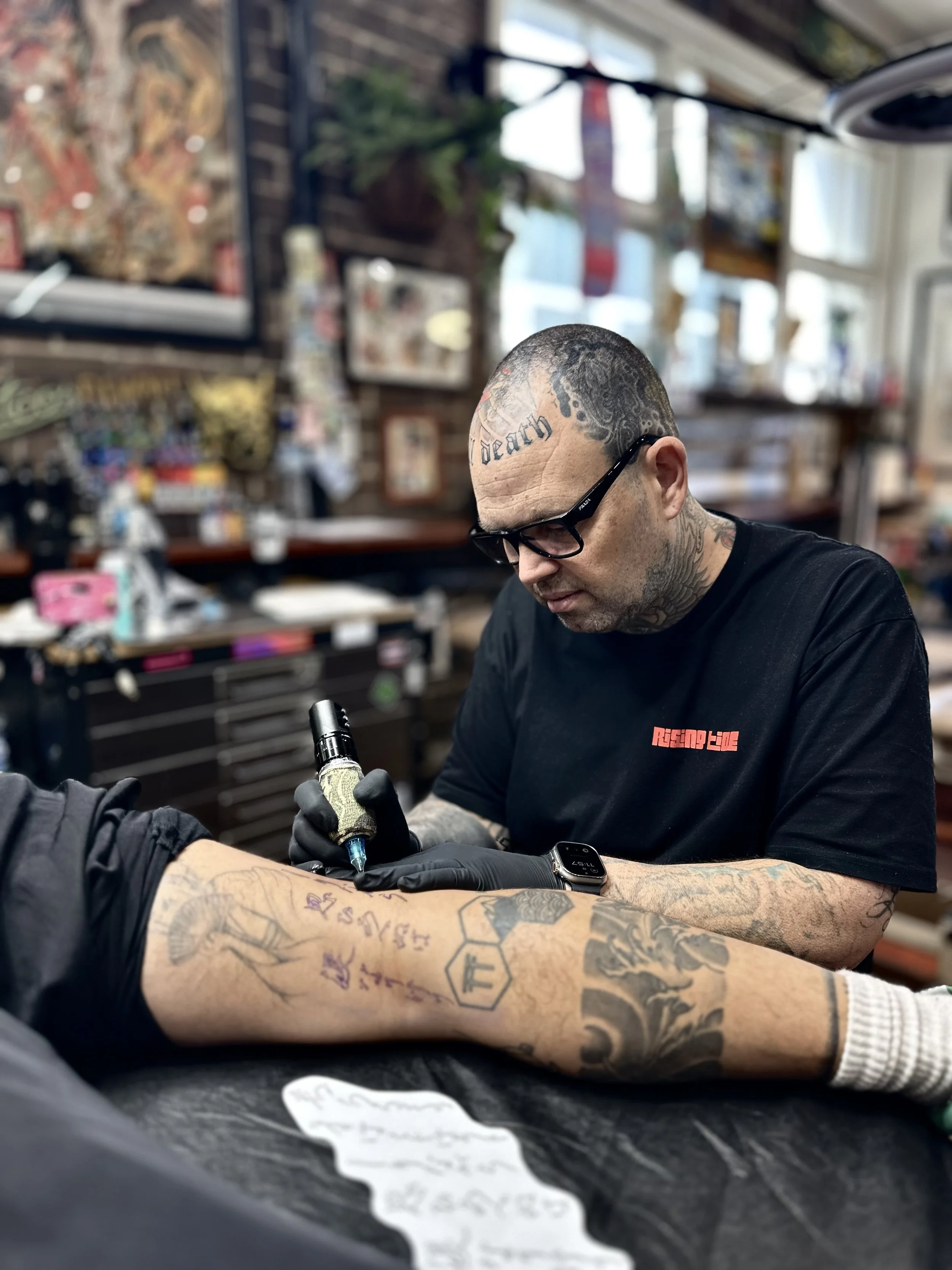 Tattoo artist wearing glasses and gloves tattoos a person's arm with a tattoo machine in a tattoo studio.