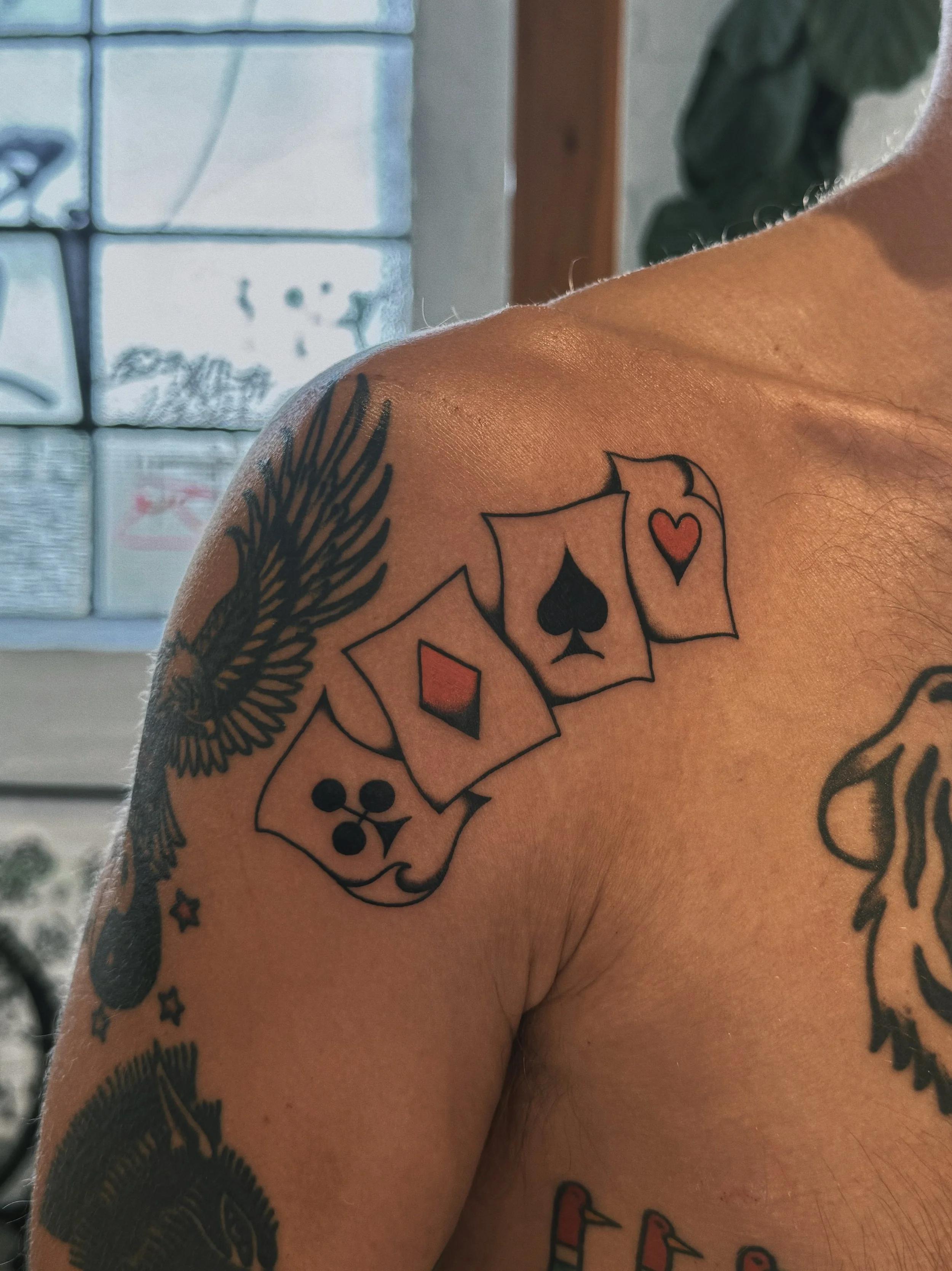 Tattoo on person's shoulder featuring four playing cards with suits of spade, heart, diamond, and club, and additional tattoos including a winged creature and various designs.
